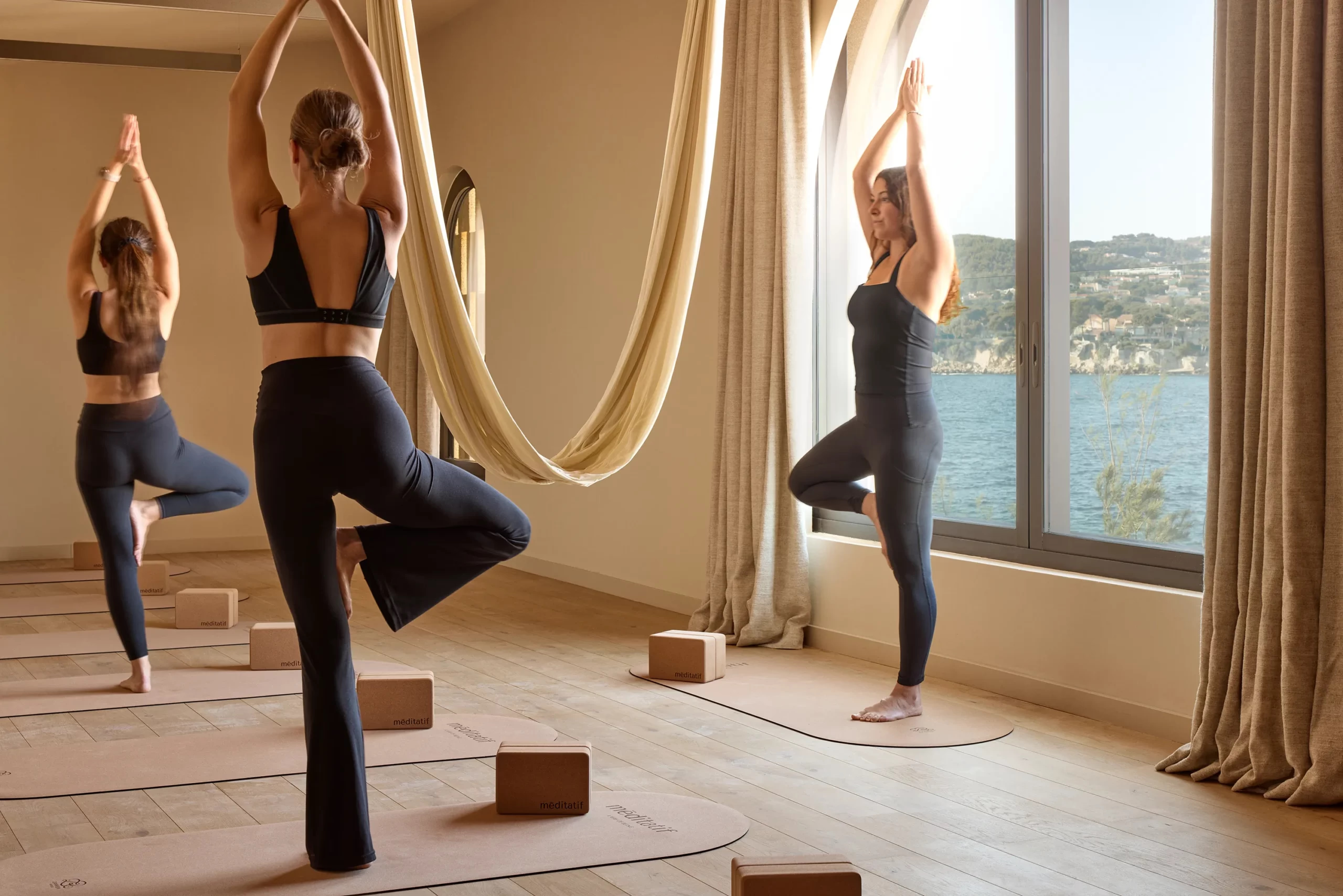 Guests hold a yoga pose on cork mats before arched windows overlooking the sea