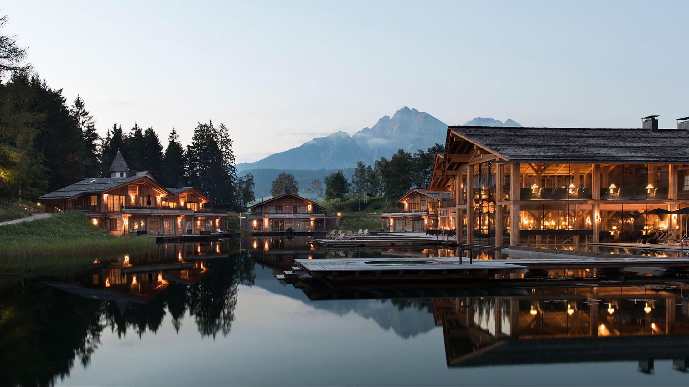 The hotel reflects in the lake at dusk, warm light glowing from wooden buildings and mountains in the distance