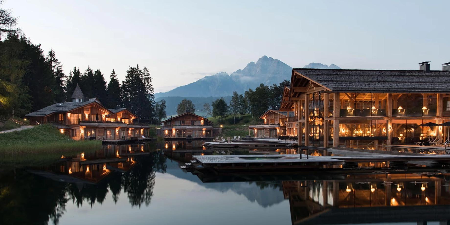 The hotel reflects in the lake at dusk, warm light glowing from wooden buildings and mountains in the distance