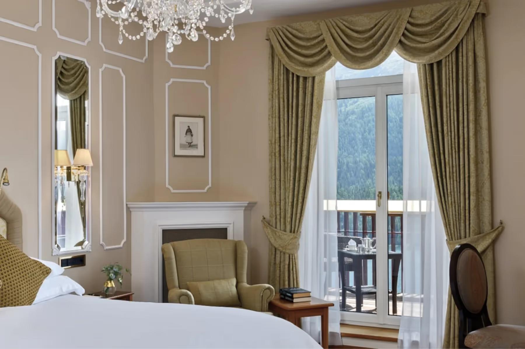 Badrutt’s Palace Hotel: suite with upholstered headboard, gold drapes, and full windows overlooking mountains