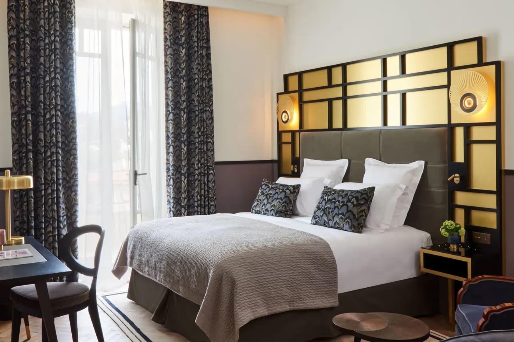A bedroom with brass headboard and geometric patterned bedding, windows with coastal views