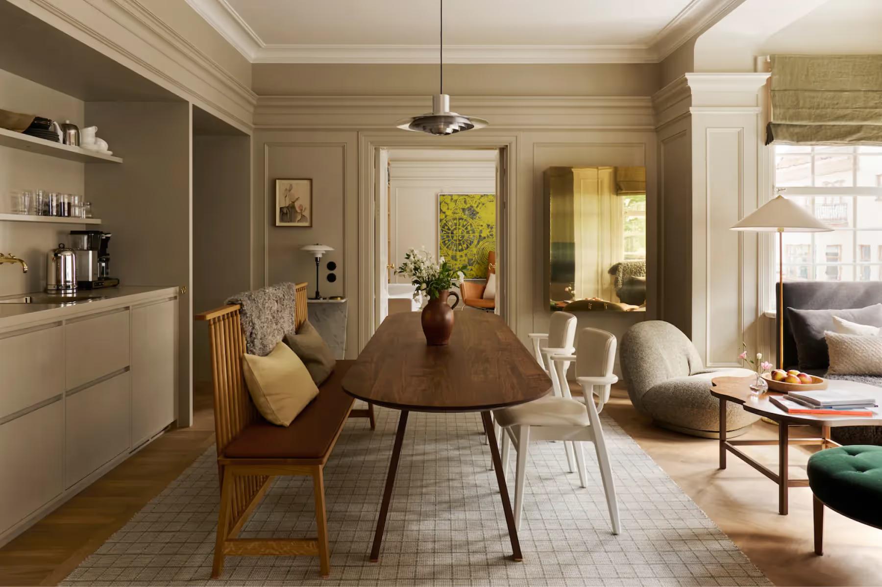 Ett Hem: a suite with dining table, wooden chairs, bookshelves, and artwork on taupe walls with bright windows