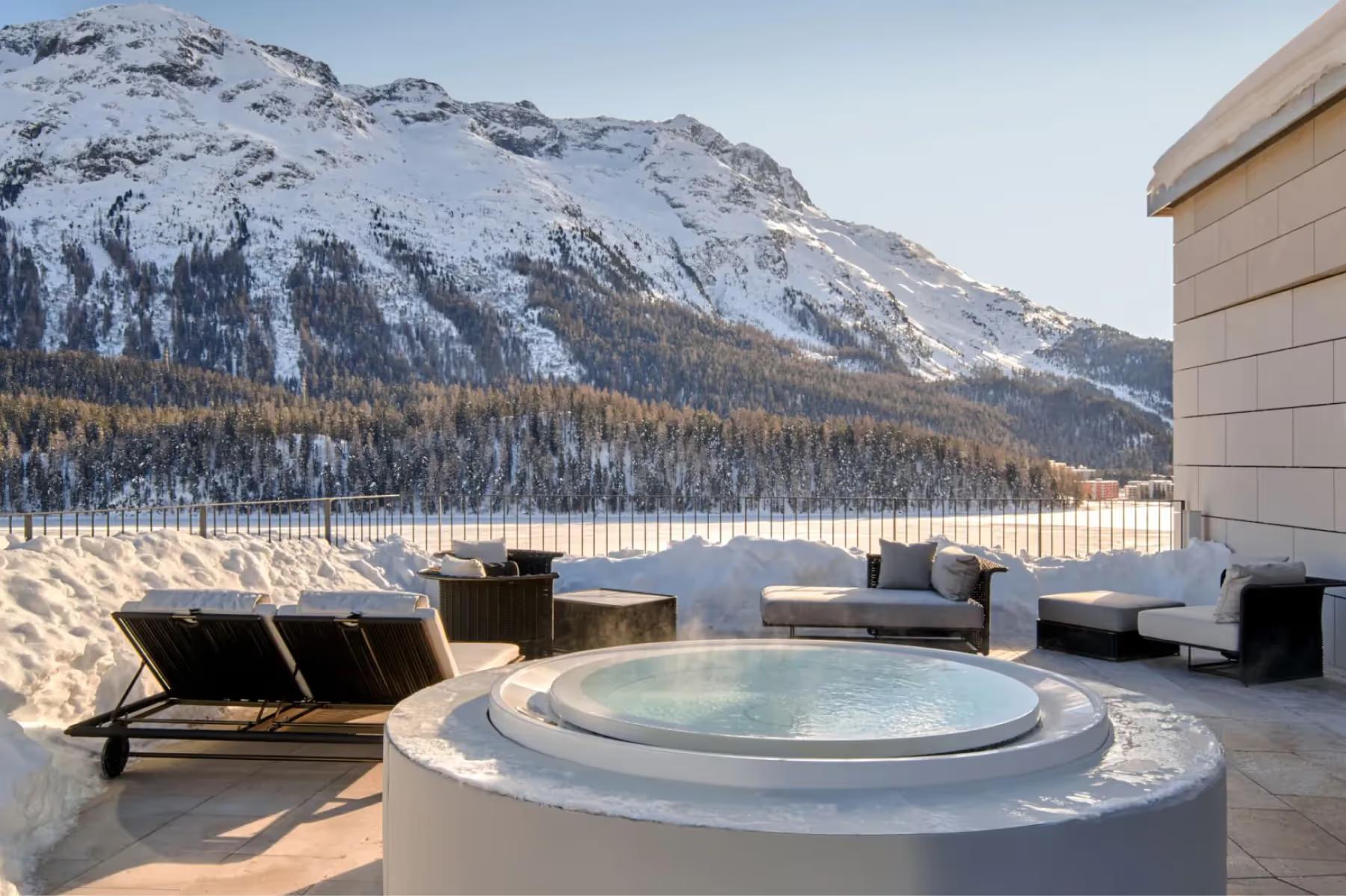 Outdoor hot tub on mountain terrace with alpine peaks and forested slopes behind
