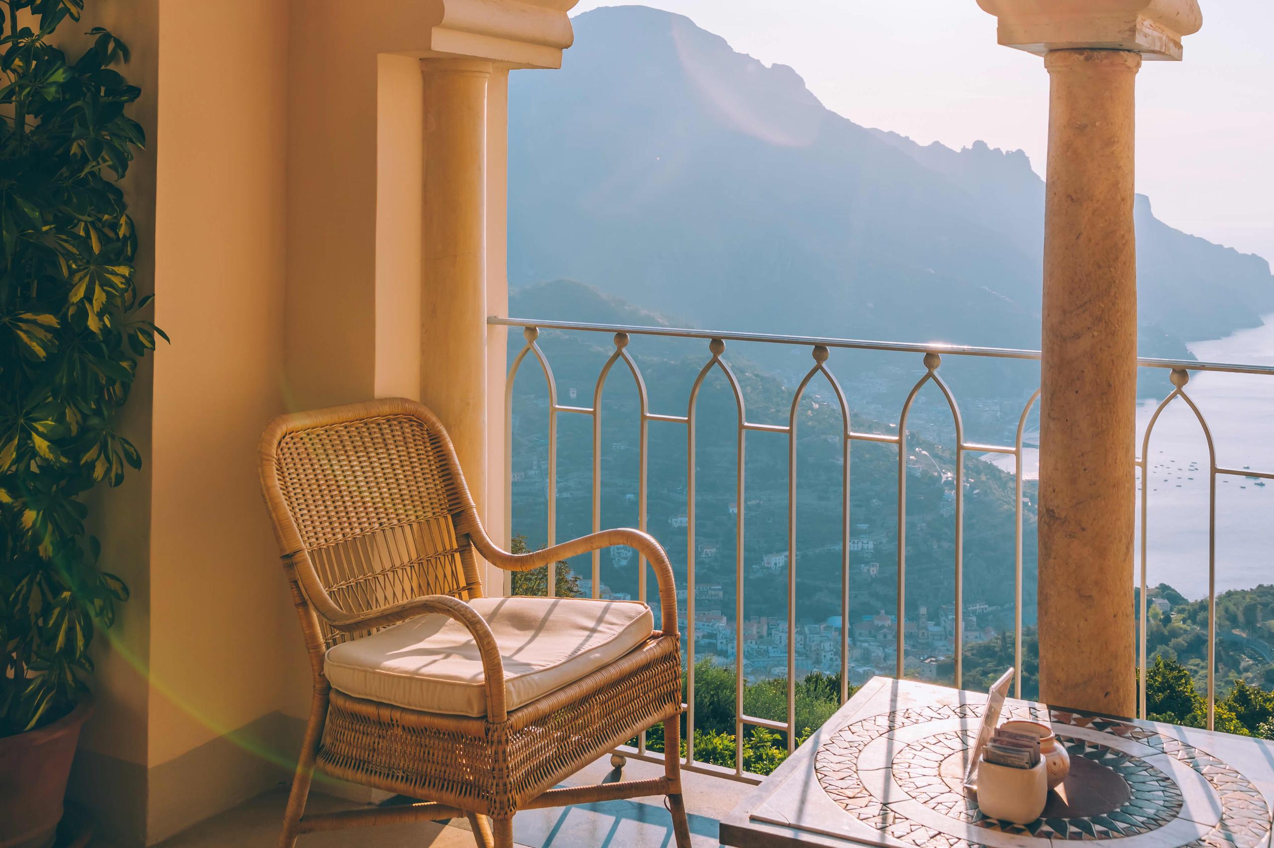 Caruso: wicker lounge chair on cream terrace with mountains, sea, and potted plants in golden light
