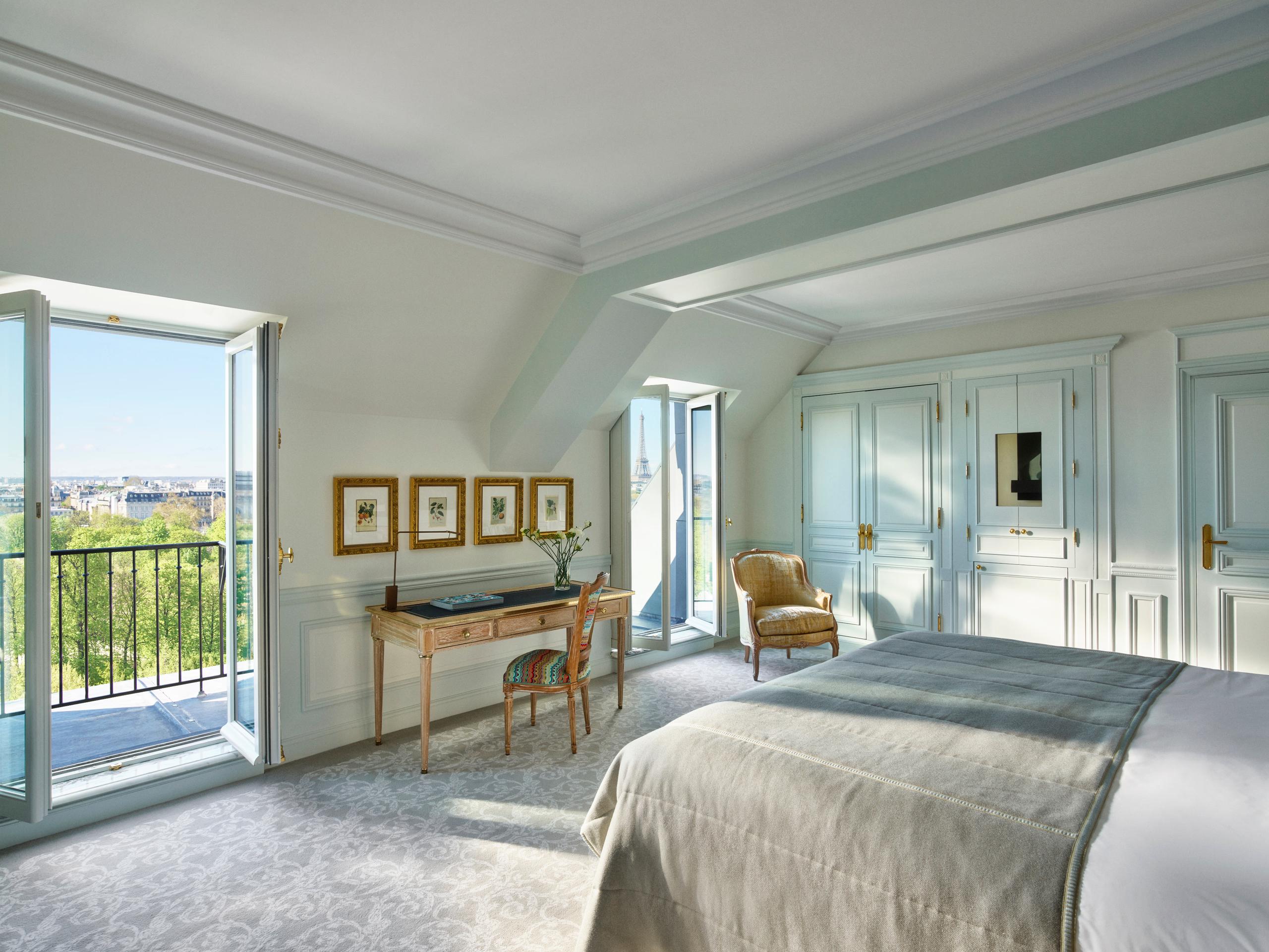 A bedroom with pale walls, queen bed, and doors opening onto a blue-railed Parisian balcony