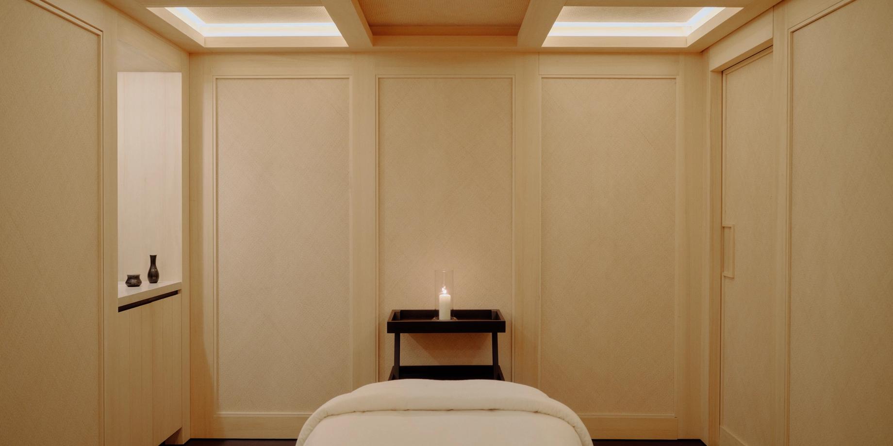 A serene Aman treatment room in pale oak, a candle on a low table below a backlit coffered ceiling