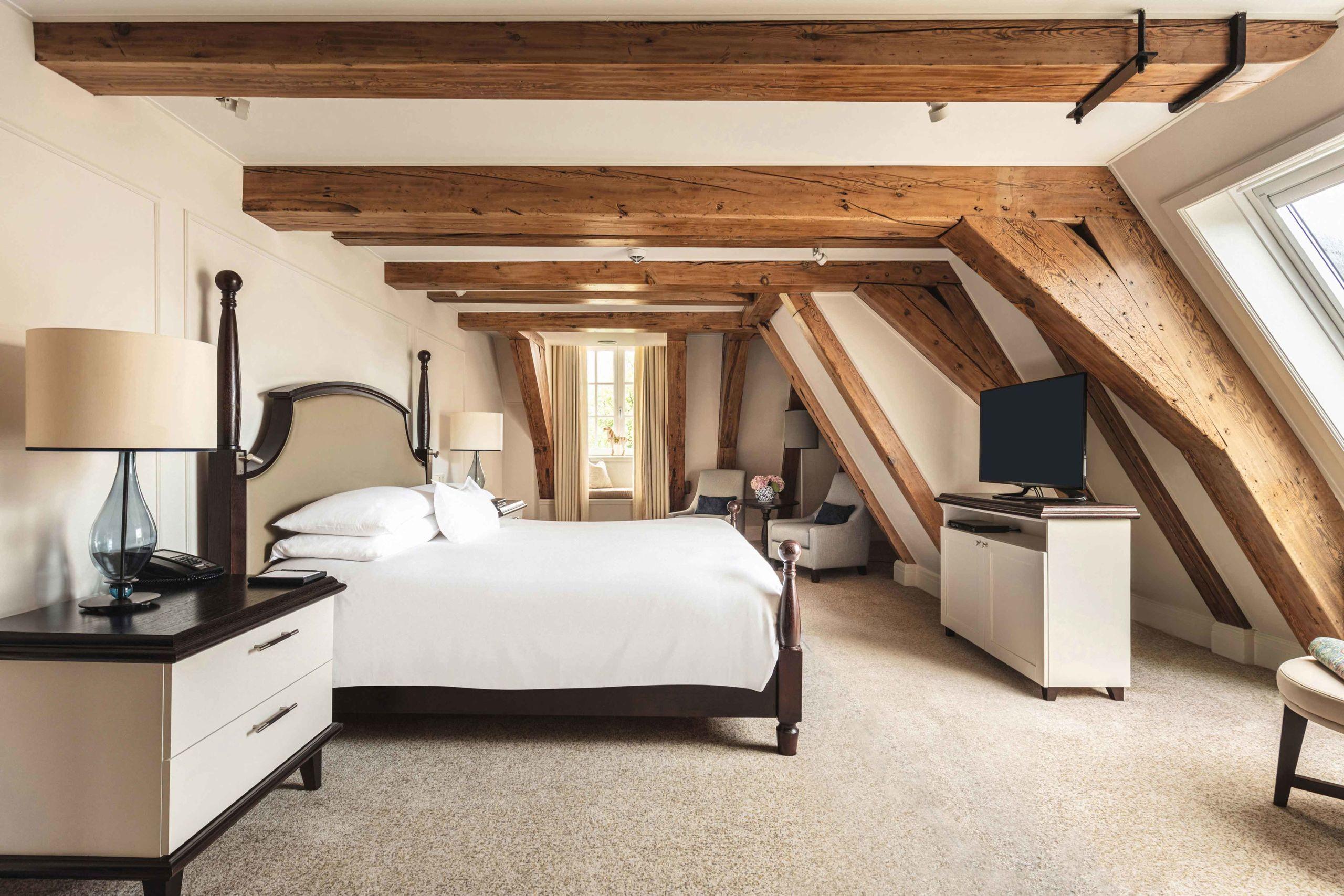 An attic bedroom with exposed timber beams, sloped ceilings, a black platform bed, and soft natural light