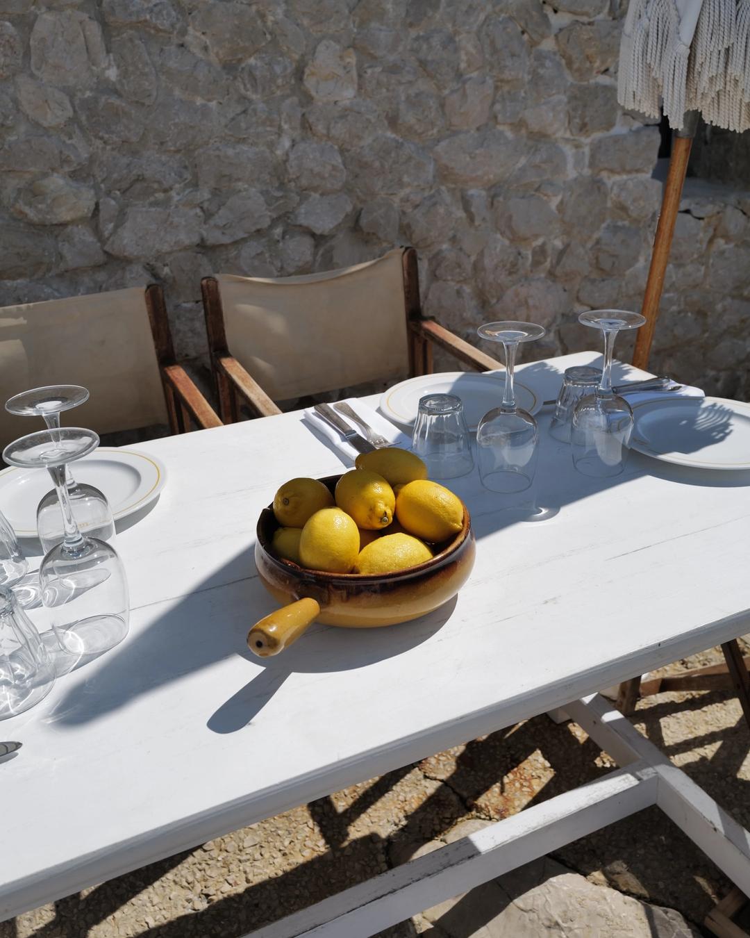 A bowl of lemons on a white table set for lunch against a dry stone wall