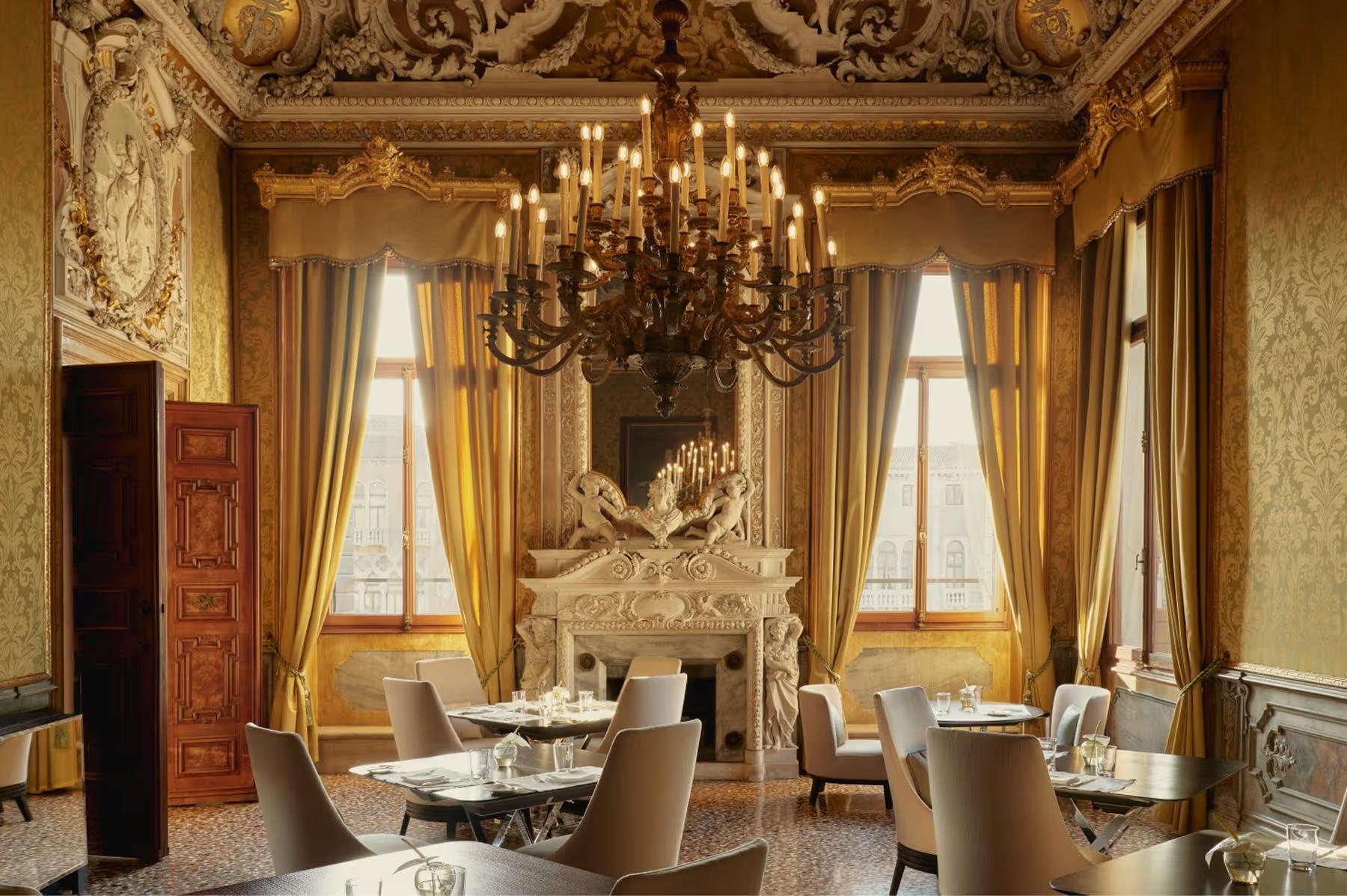 Aman Venice: a formal dining room with ornate gilded walls, crystal chandelier, draped windows, and round tables with chairs