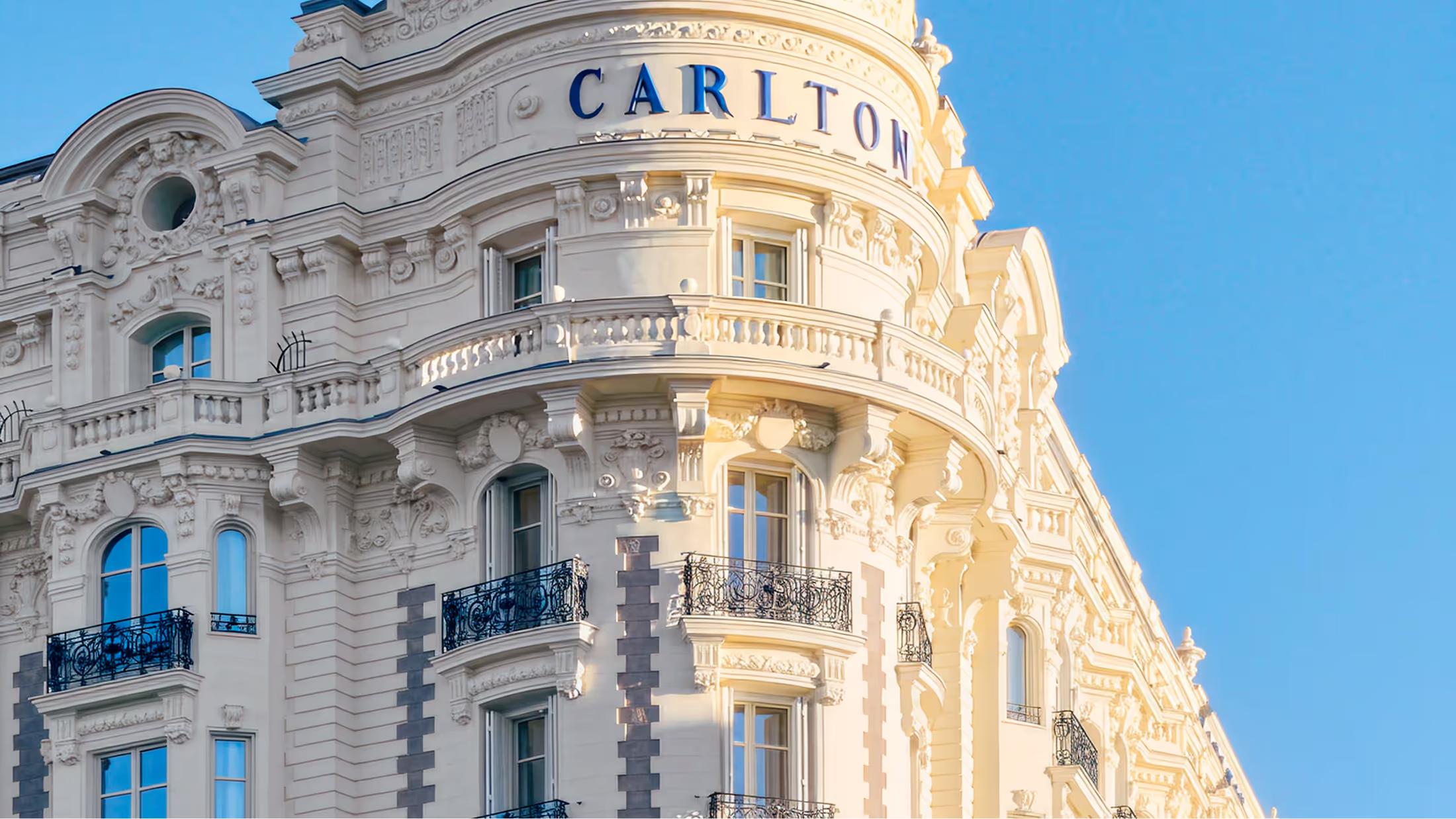 The curved cream stone facade of Carlton Cannes with its iconic turret dome under clear blue sky