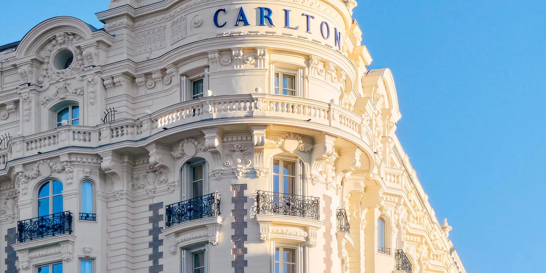 The curved cream stone facade of Carlton Cannes with its iconic turret dome under clear blue sky