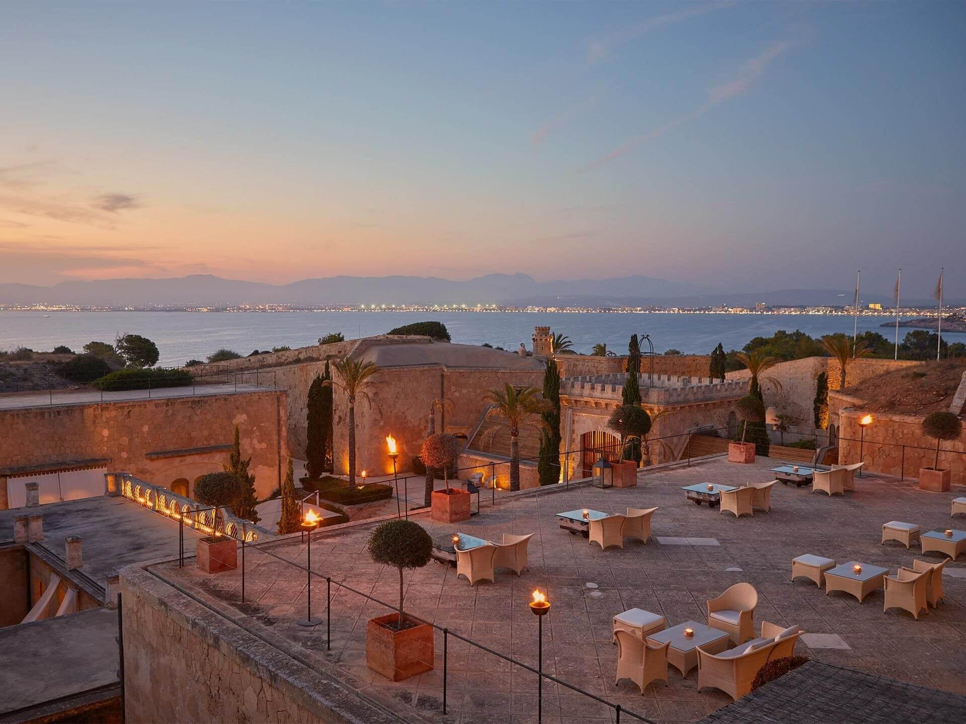 A rooftop terrace at twilight with cream loungers, lit lanterns, and sea horizon