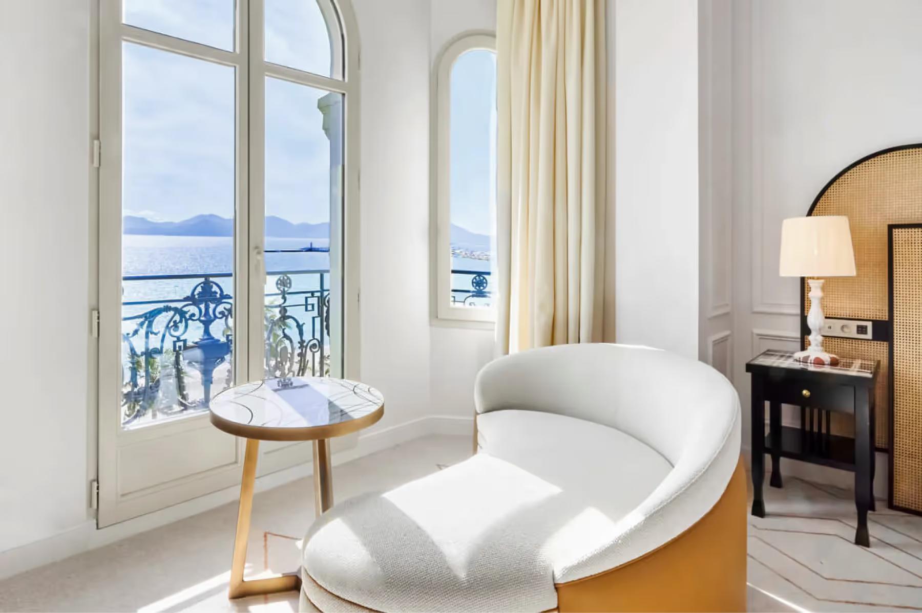 Seating nook with curved white lounge chair by windows overlooking the Mediterranean with a small brass-legged table