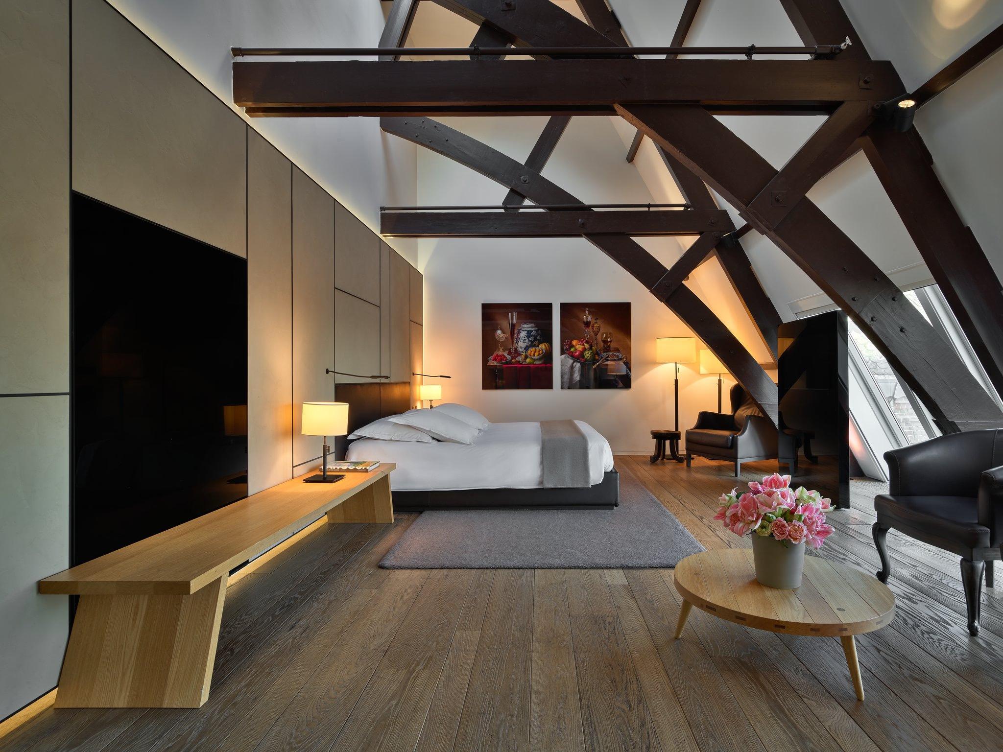Mandarin Oriental Conservatorium, Amsterdam: loft bedroom under dark timber roof beams, with a wooden bench, a round table with pink flowers, and two framed still lifes above the bed