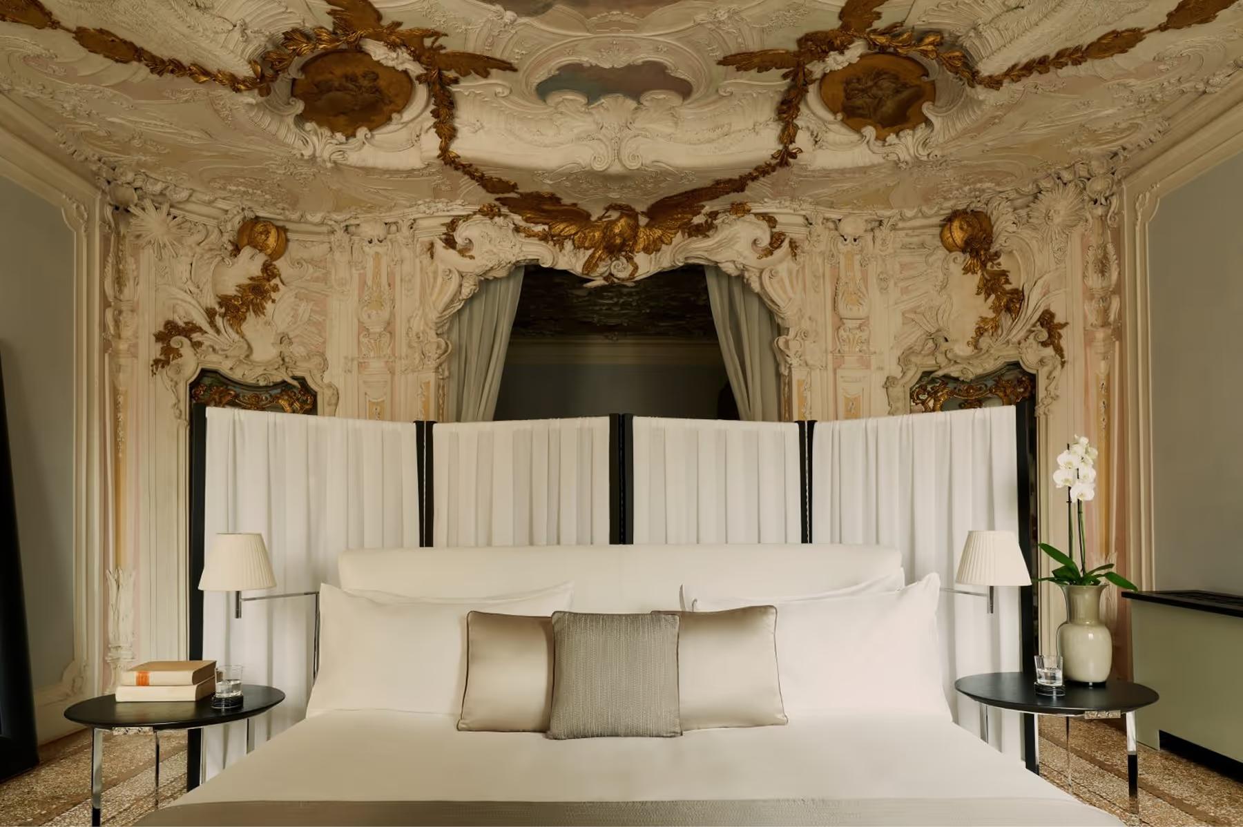 Aman Venice: a bedroom with ornate frescoed ceiling, four-poster bed with white bedding, and matching carved wooden nightstands