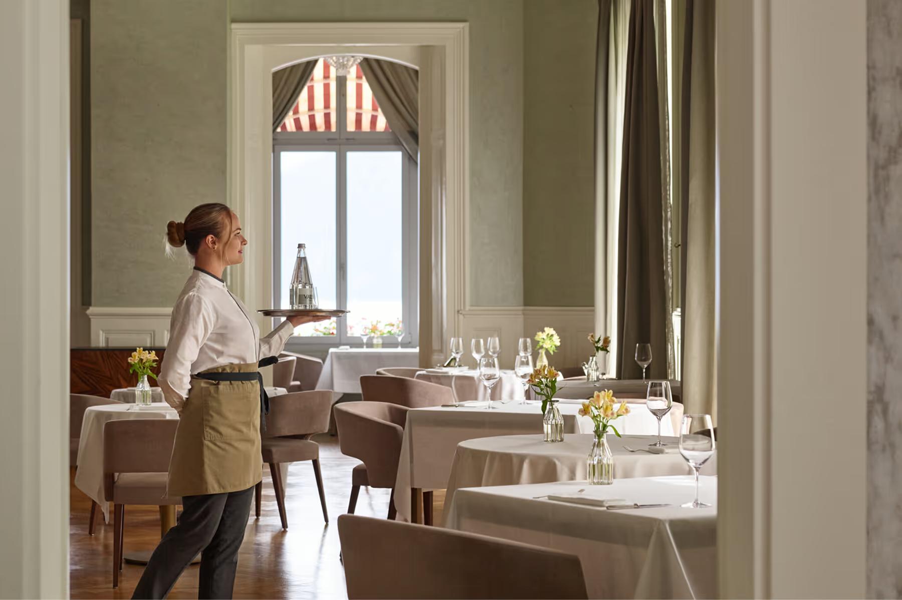 Private dining room with pale walls, server in white service dress, white-clothed tables, and tall windows