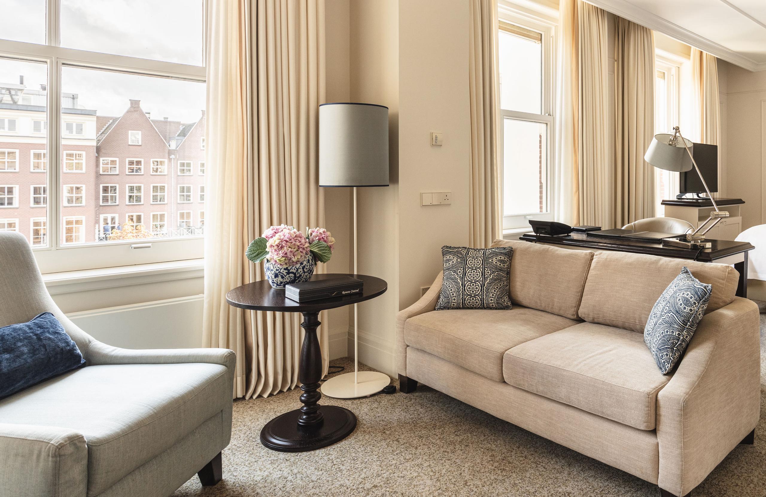 A sunlit lounge with cream armchairs and a beige sofa framing a view of 17th-century buildings across a canal