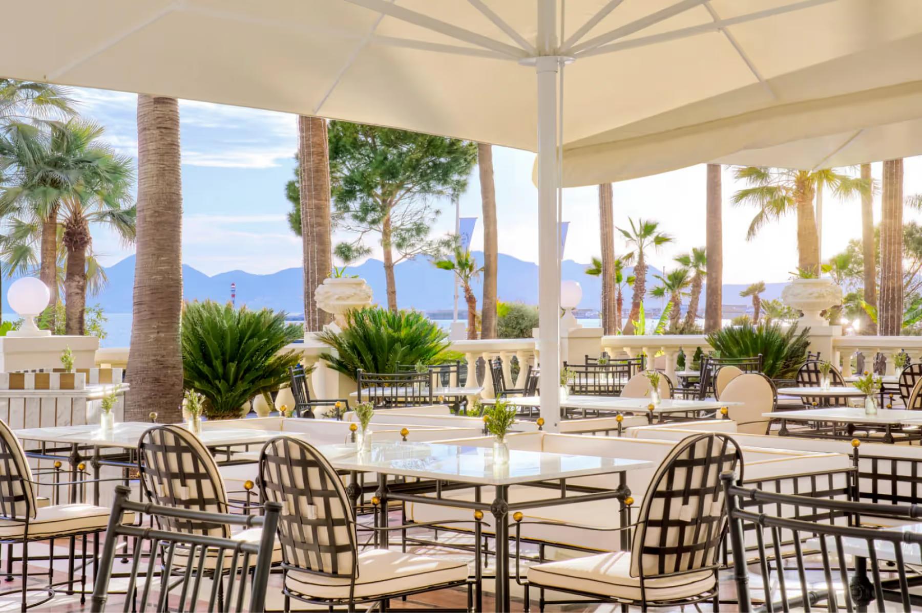 Carlton Cannes: terrace dining area under white umbrellas with sea views, palm trees, and woven dining chairs