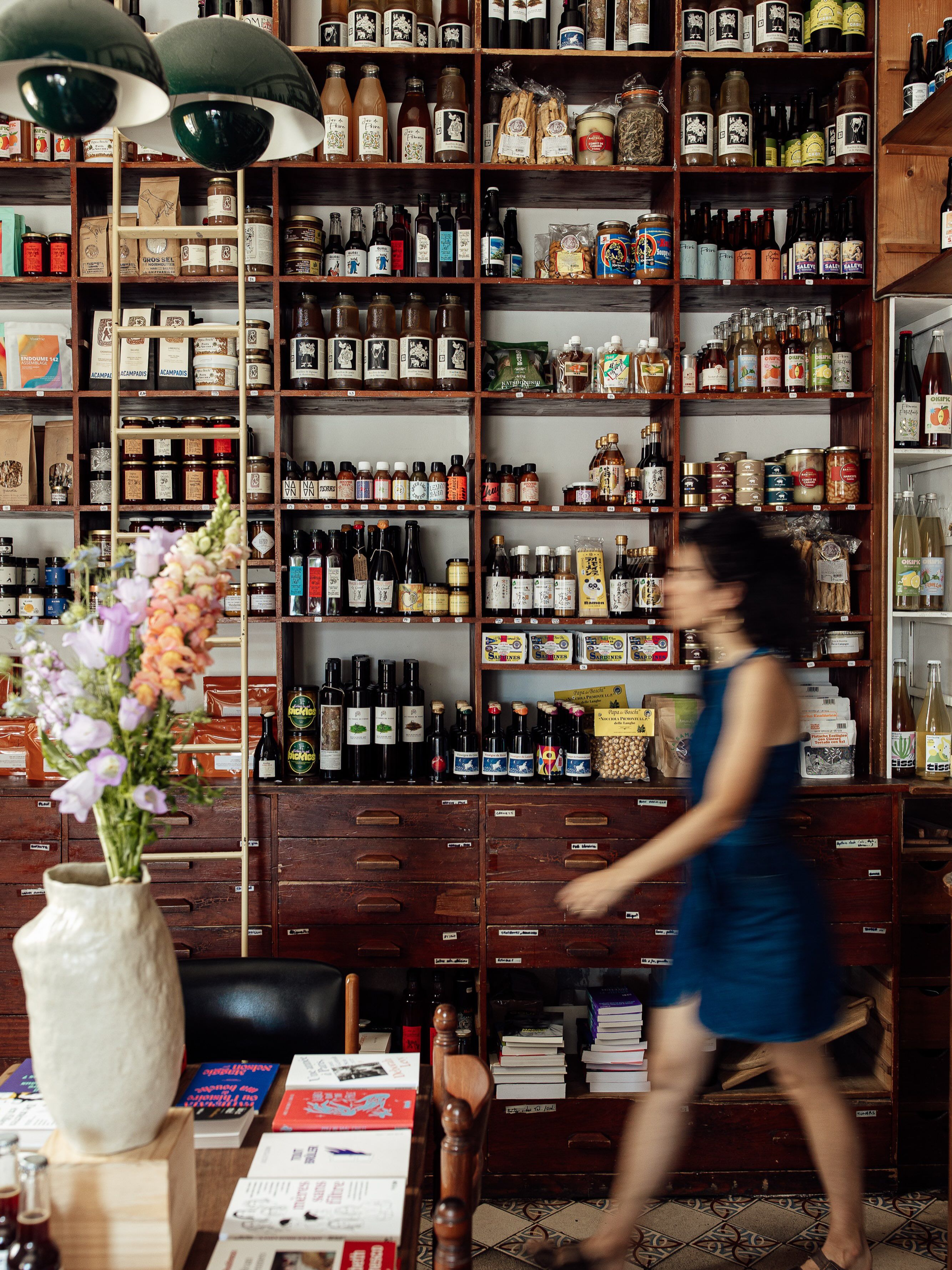 Shelves stocked with bottles and jars at Provisions, a grocery and bookshop in Marseille, with a woman walking past