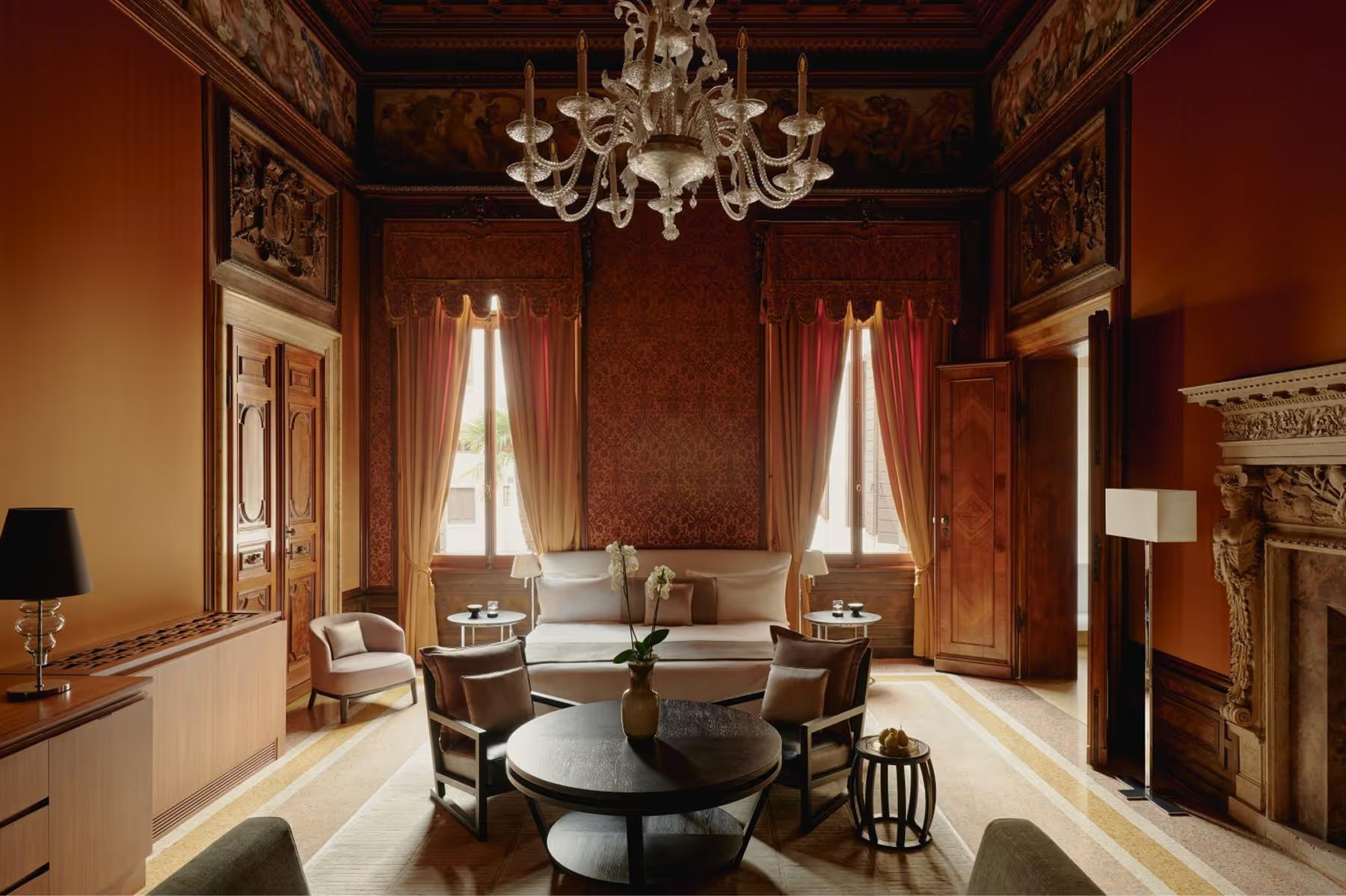 Aman Venice: a jewel-toned suite with deep red walls, ornate painted ceiling, white marble fireplace, and cream seating