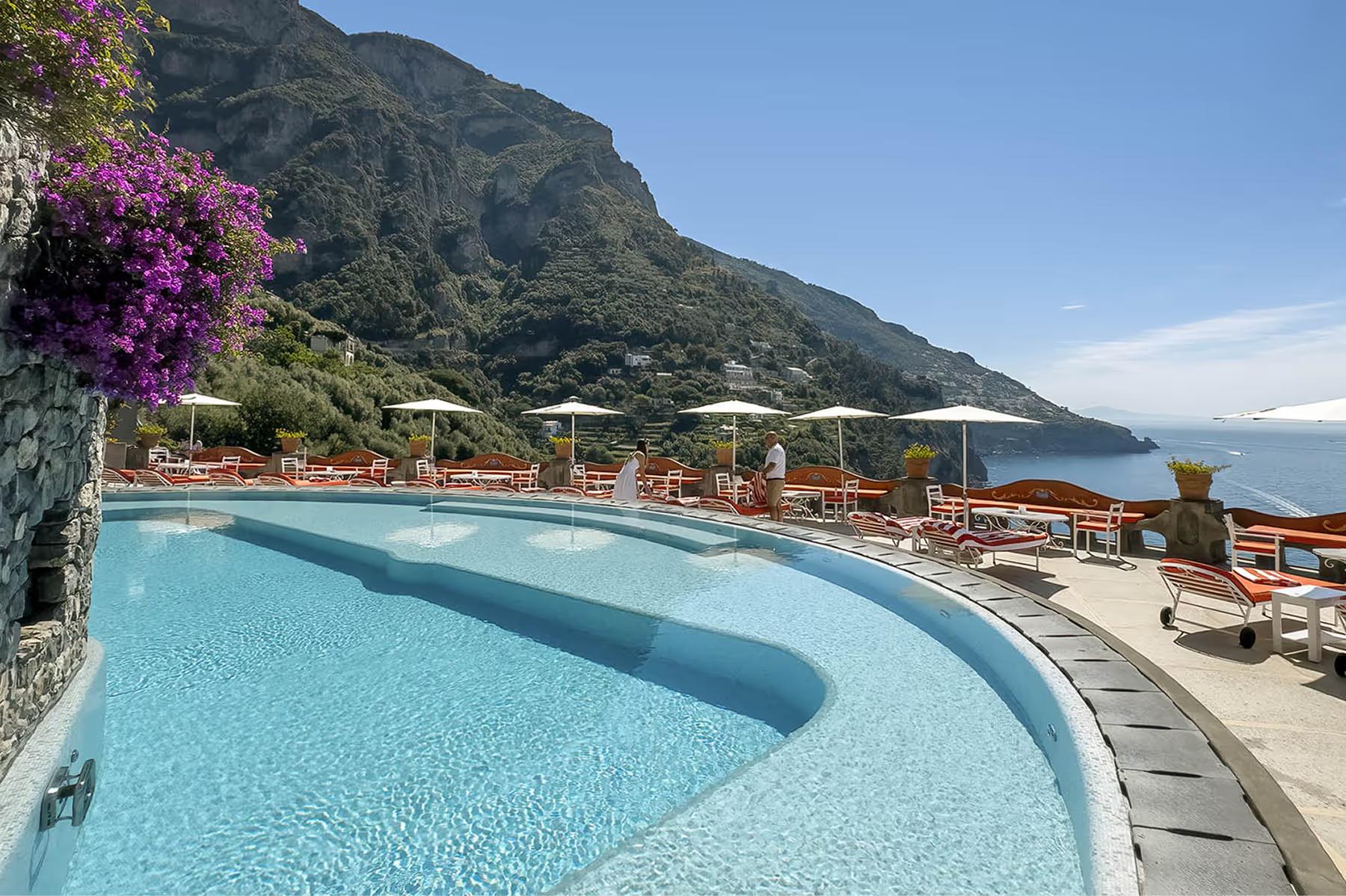 Curved infinity pool with turquoise water overlooking Mediterranean, flowering plants, and cliffside setting