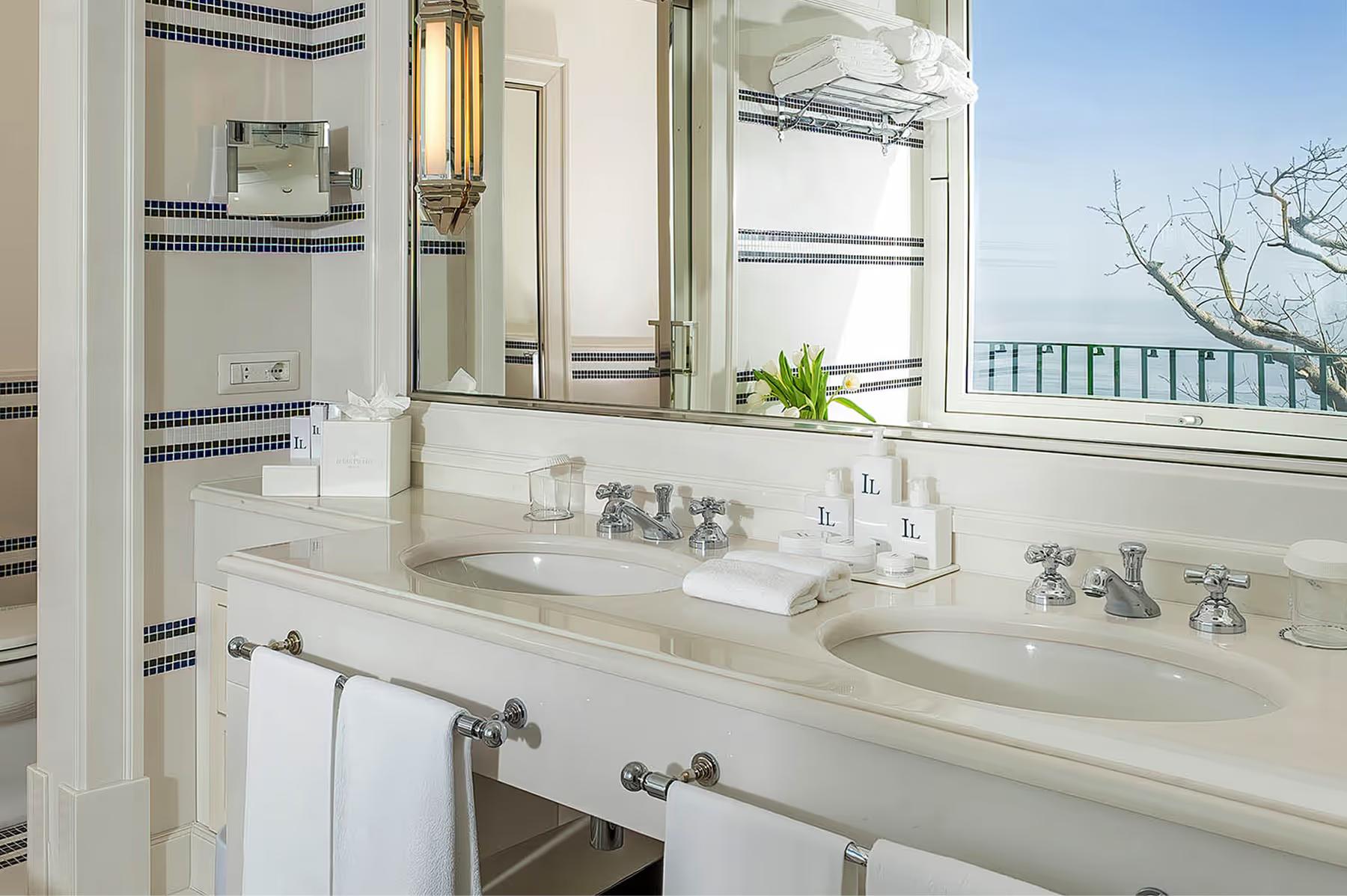 Il San Pietro di Positano: marble vanity with dual sinks, brass fixtures, windows overlooking Mediterranean, white tile surround