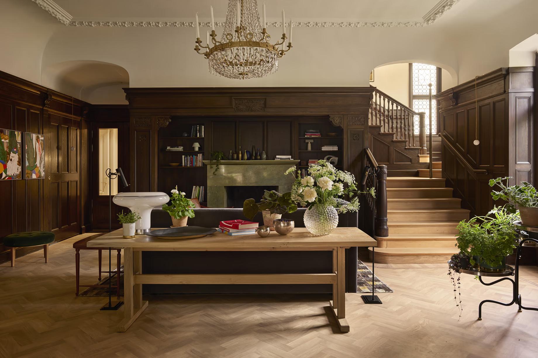 A wood-paneled hall with a crystal chandelier, a green stone fireplace, a long wooden table and a staircase