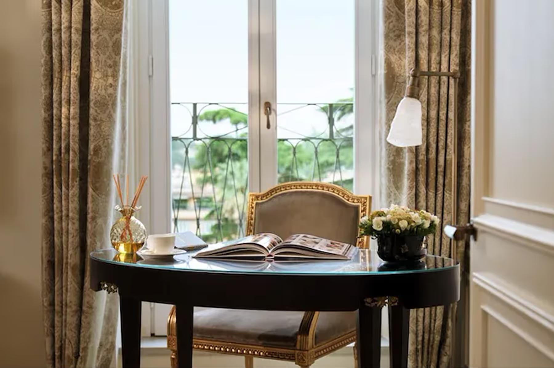 A writing desk by tall windows with gold curtains, topped with an open book and flowers overlooking the city
