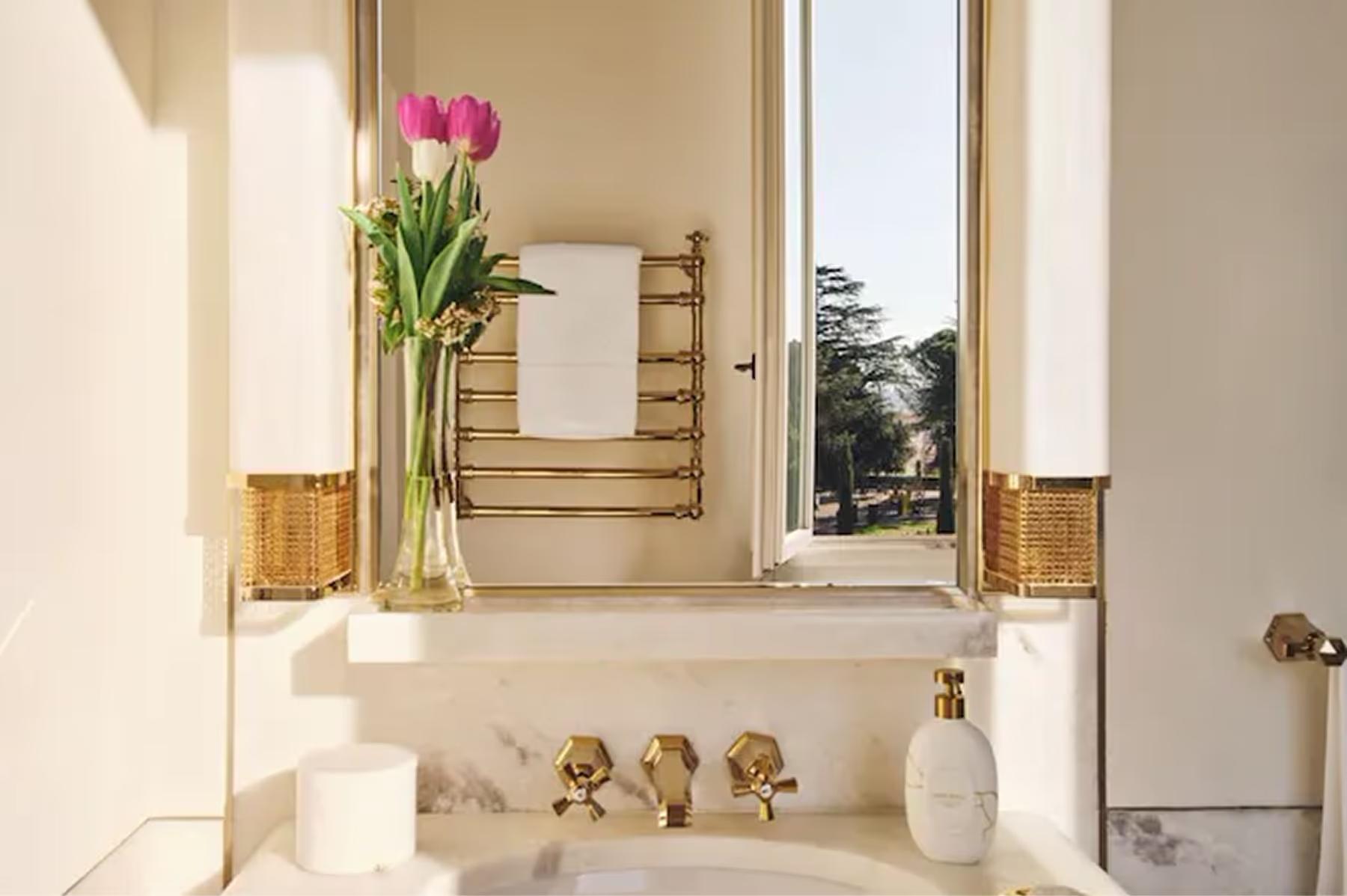 Hotel Eden: a marble bathroom with a vanity, brass fixtures, fresh flowers in a vase, and natural light from a window