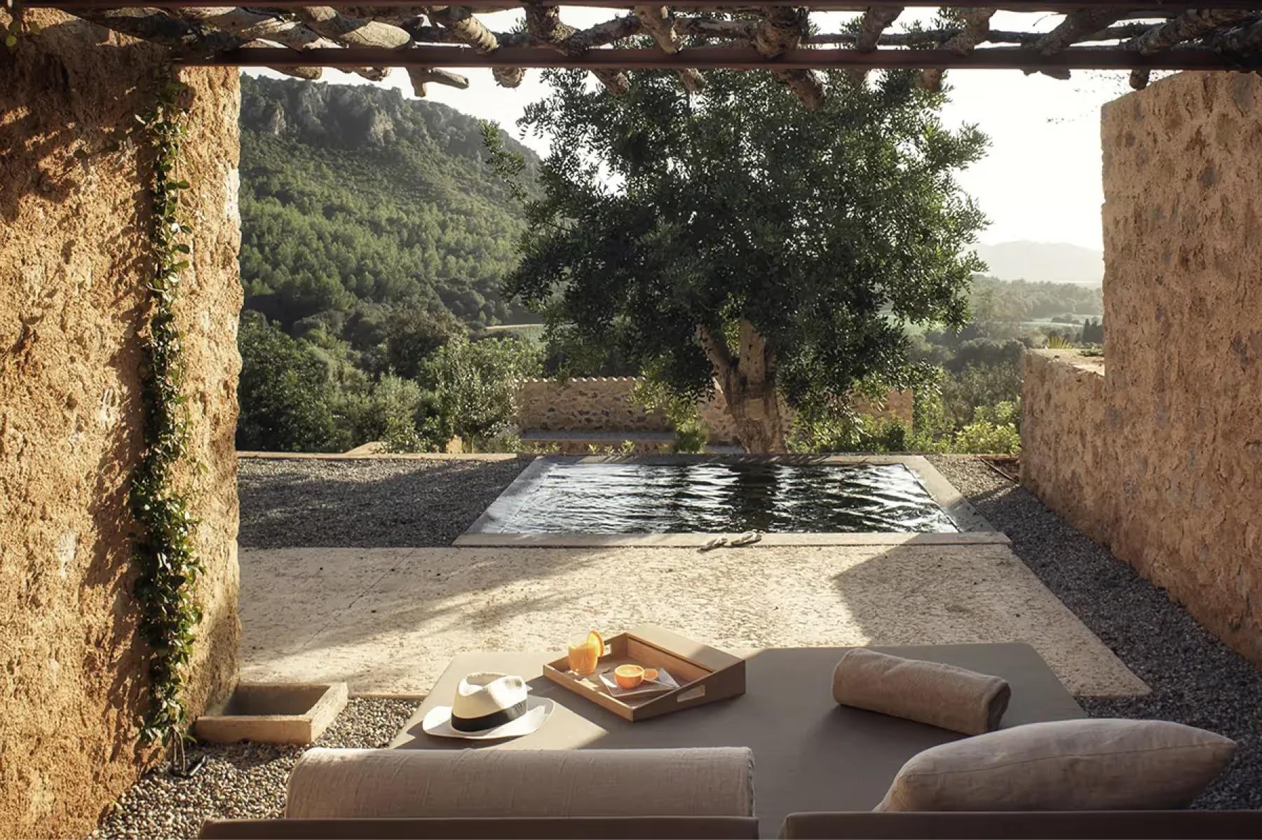 Covered lounge area framing a blue-toned pool, stone walls, and distant Mediterranean landscape views