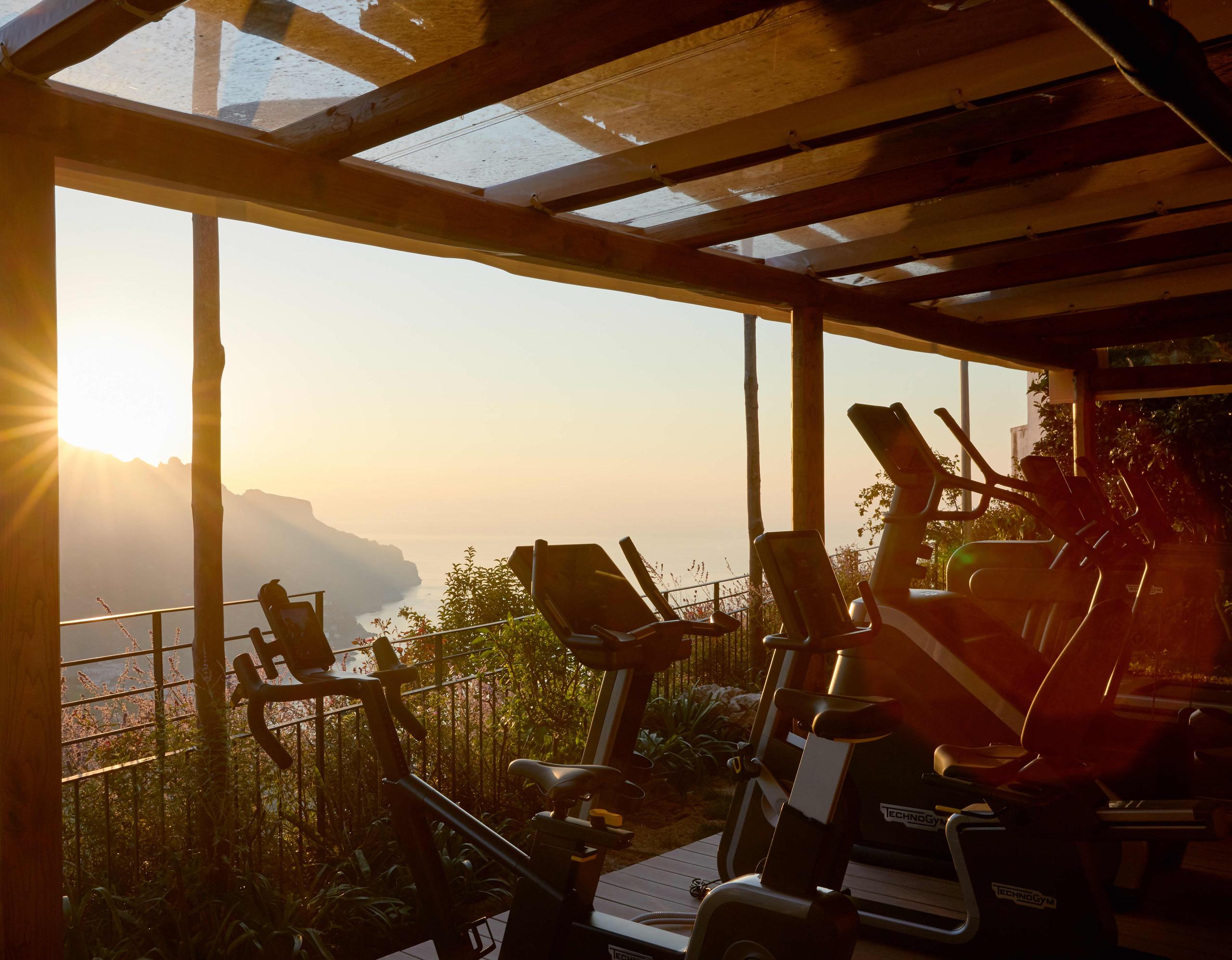 Caruso: fitness room overlooking coast at sunset with exercise equipment and mountain silhouettes