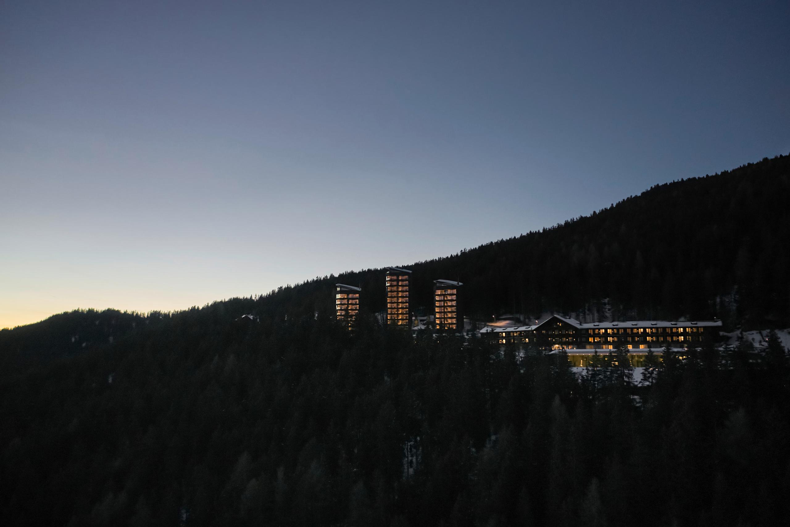 The hotel facade glowing warmly at dusk against a mountainside forest landscape