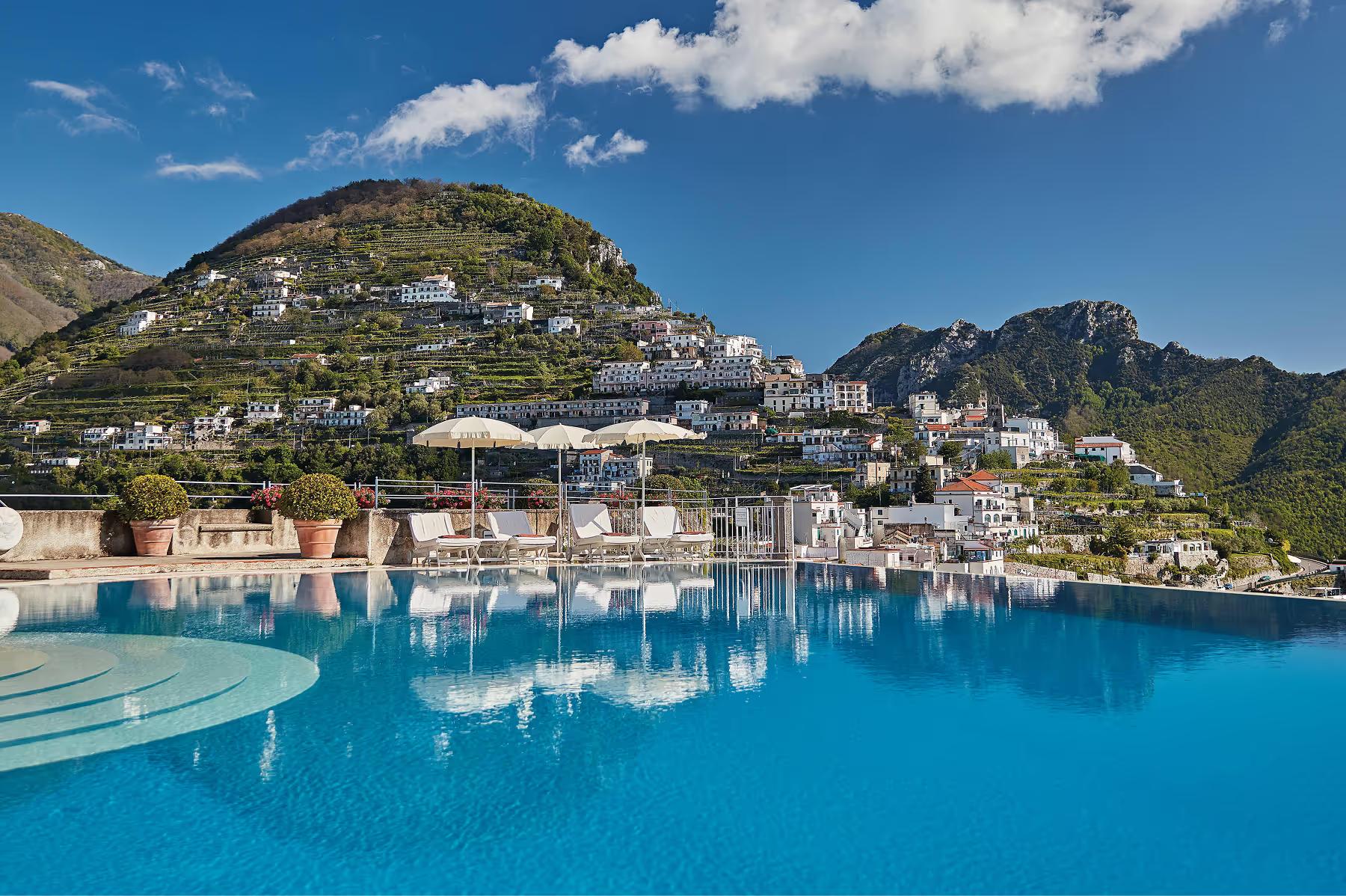 Aerial view of turquoise infinity pool reflecting mountainous hilltop village and coastal scenery