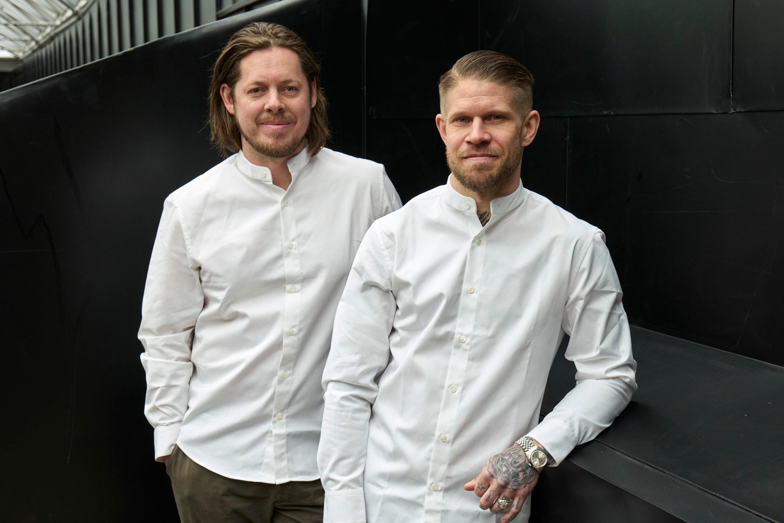 Two chefs in white chef coats standing before their restaurant kitchen