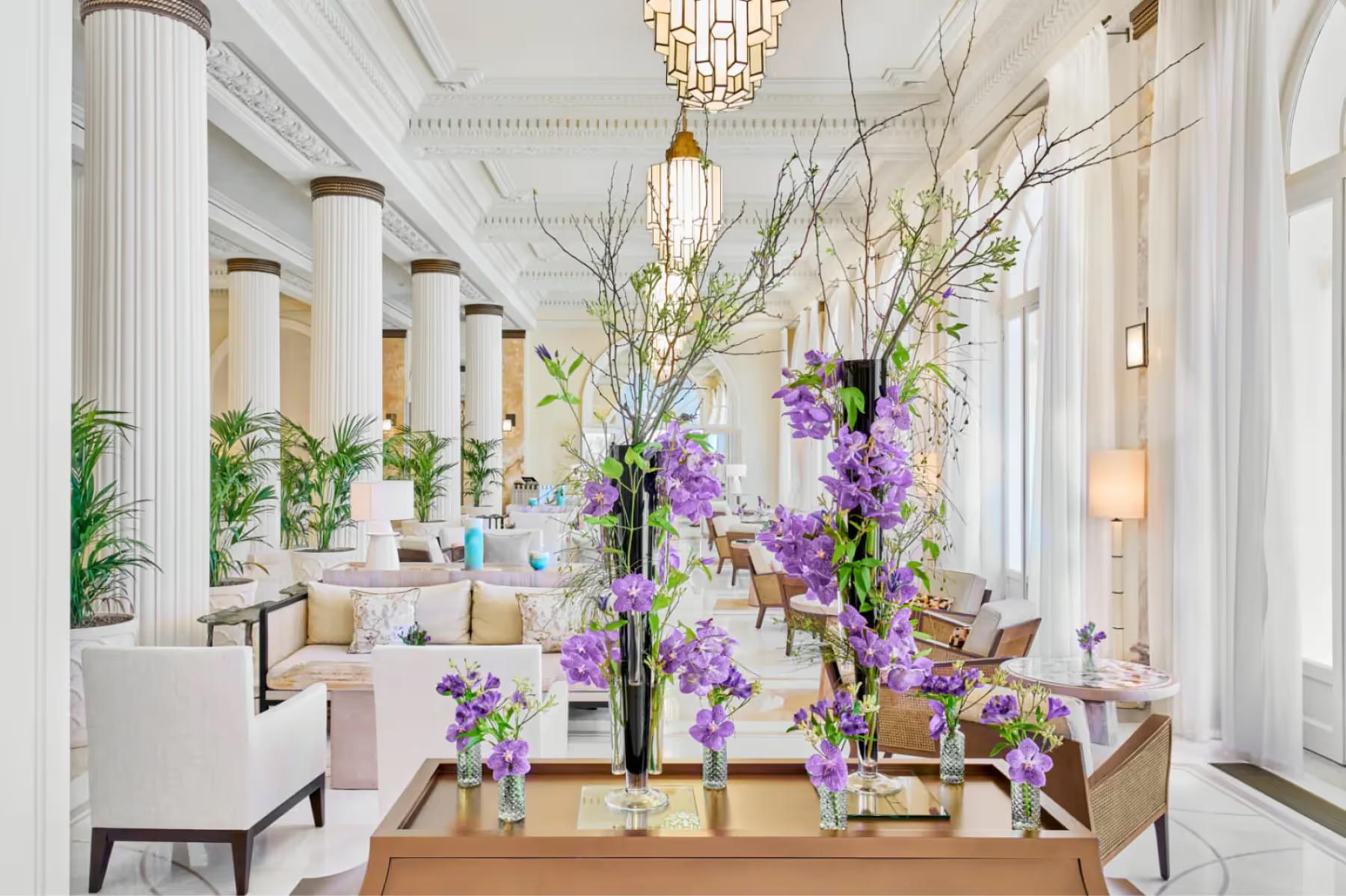 Carlton Cannes: bright lobby with tall white columns, purple flower arrangements on a wooden table, and chandelier lighting