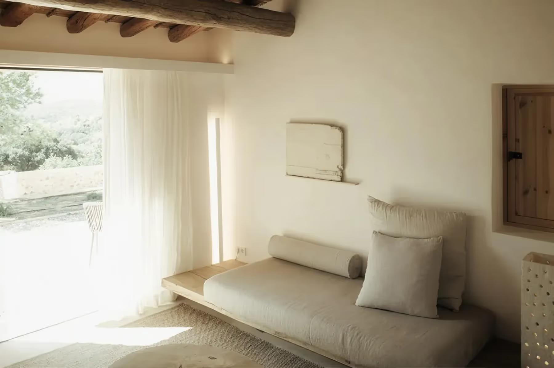 Es Racó d’Artà: calm residential alcove with daybed, cushions, and tall window providing soft natural light and garden view