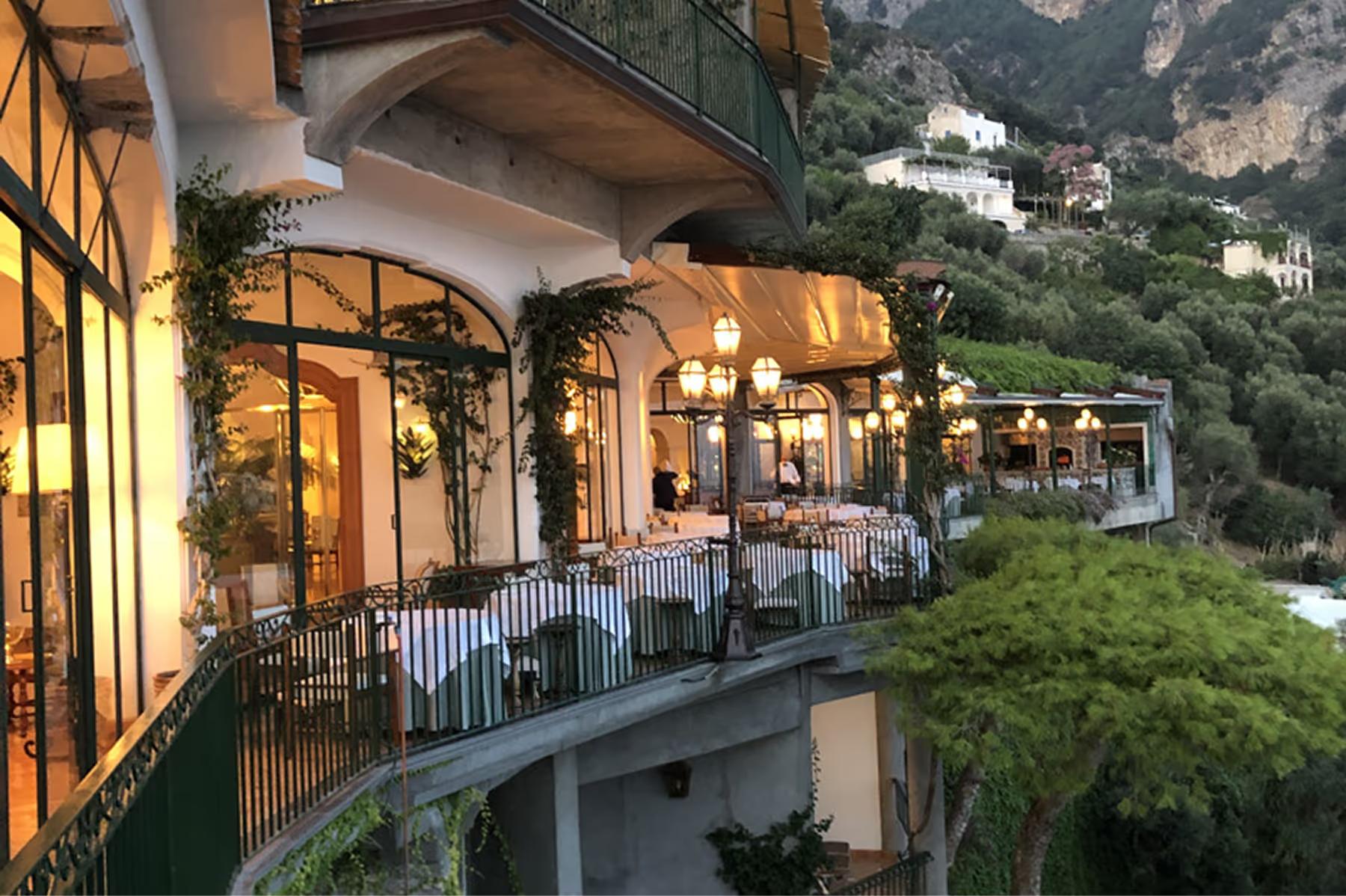 Il San Pietro di Positano: evening-lit restaurant terrace with warm lighting, railings, and views across coastal town below