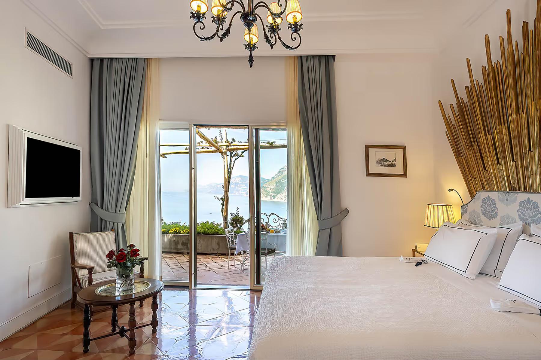 Il San Pietro di Positano: bedroom suite with bed, chandelier, terrace doors, wood flooring, and glimpse of coastal landscape