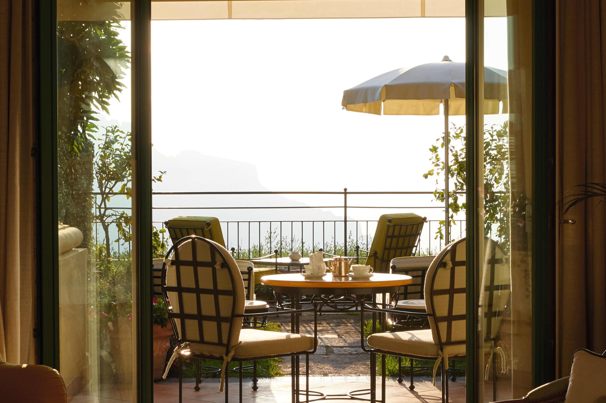 Caruso: terrace view to sea framed by open wood doors, round table with yellow umbrella, wicker chairs in afternoon light
