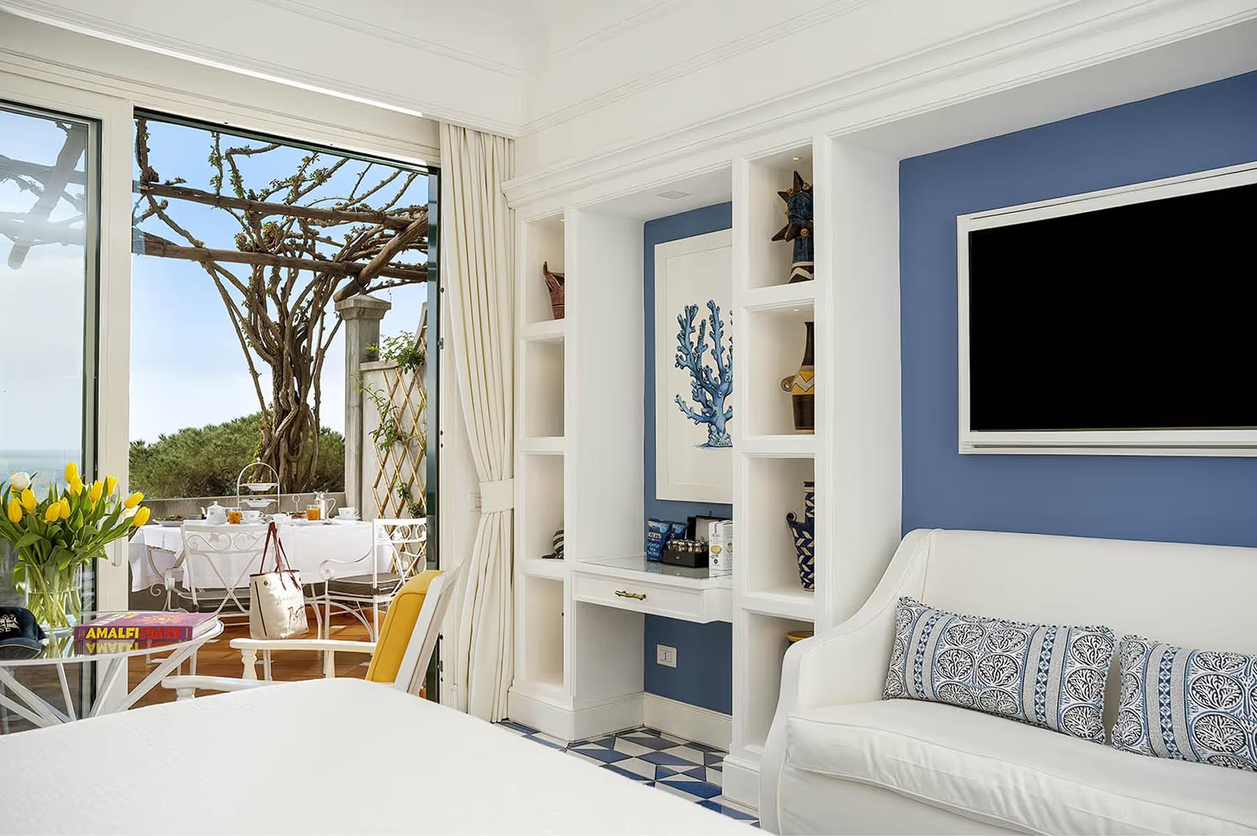 Il San Pietro di Positano: bedroom with white linens, built-in shelving, floor-to-ceiling windows, sea view terrace, and blue accent wall