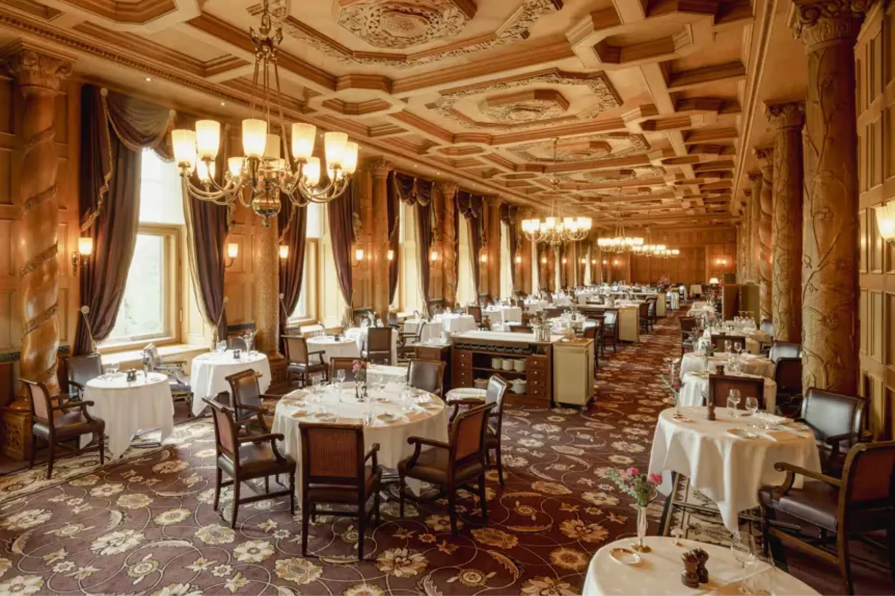 Suvretta House: an elegant dining room with ornate gold ceiling, brass wall sconces, and white linens on every table