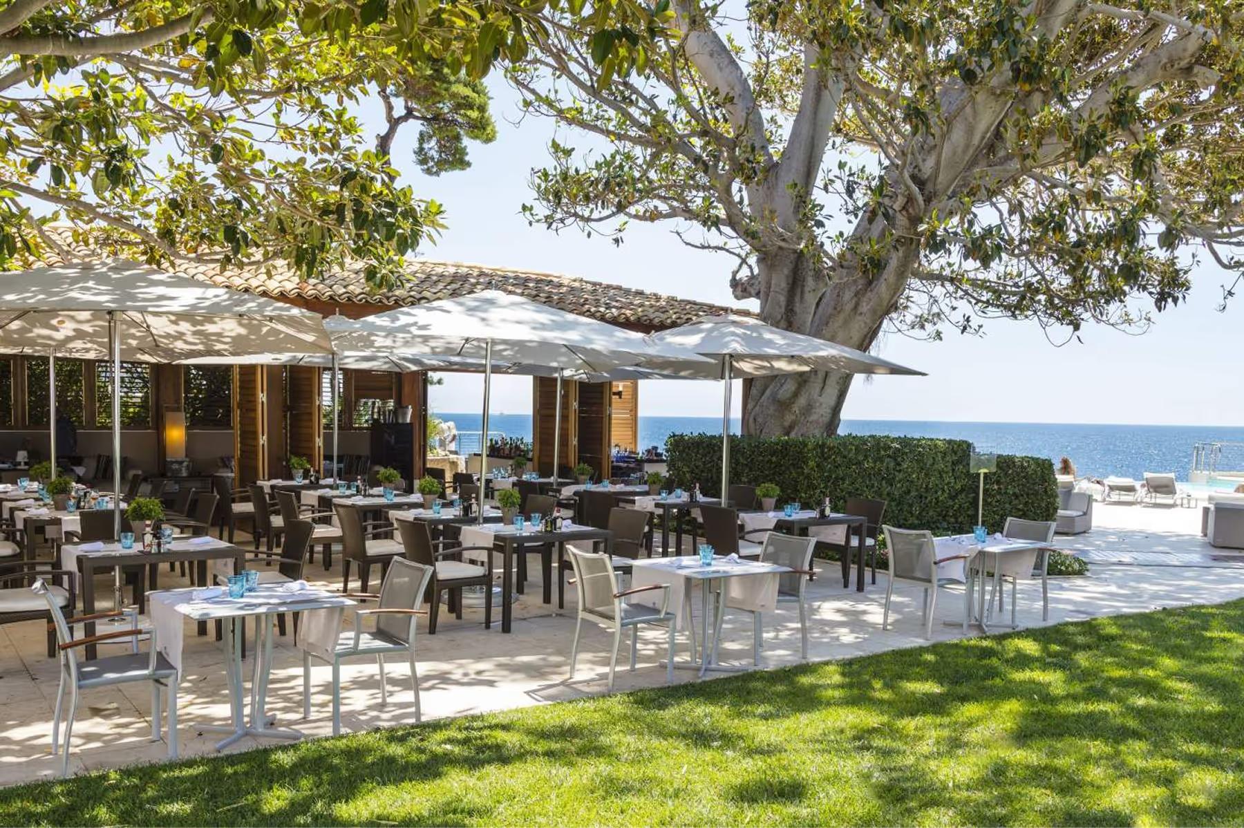 Dining tables arranged under shaded structures with Mediterranean seascape