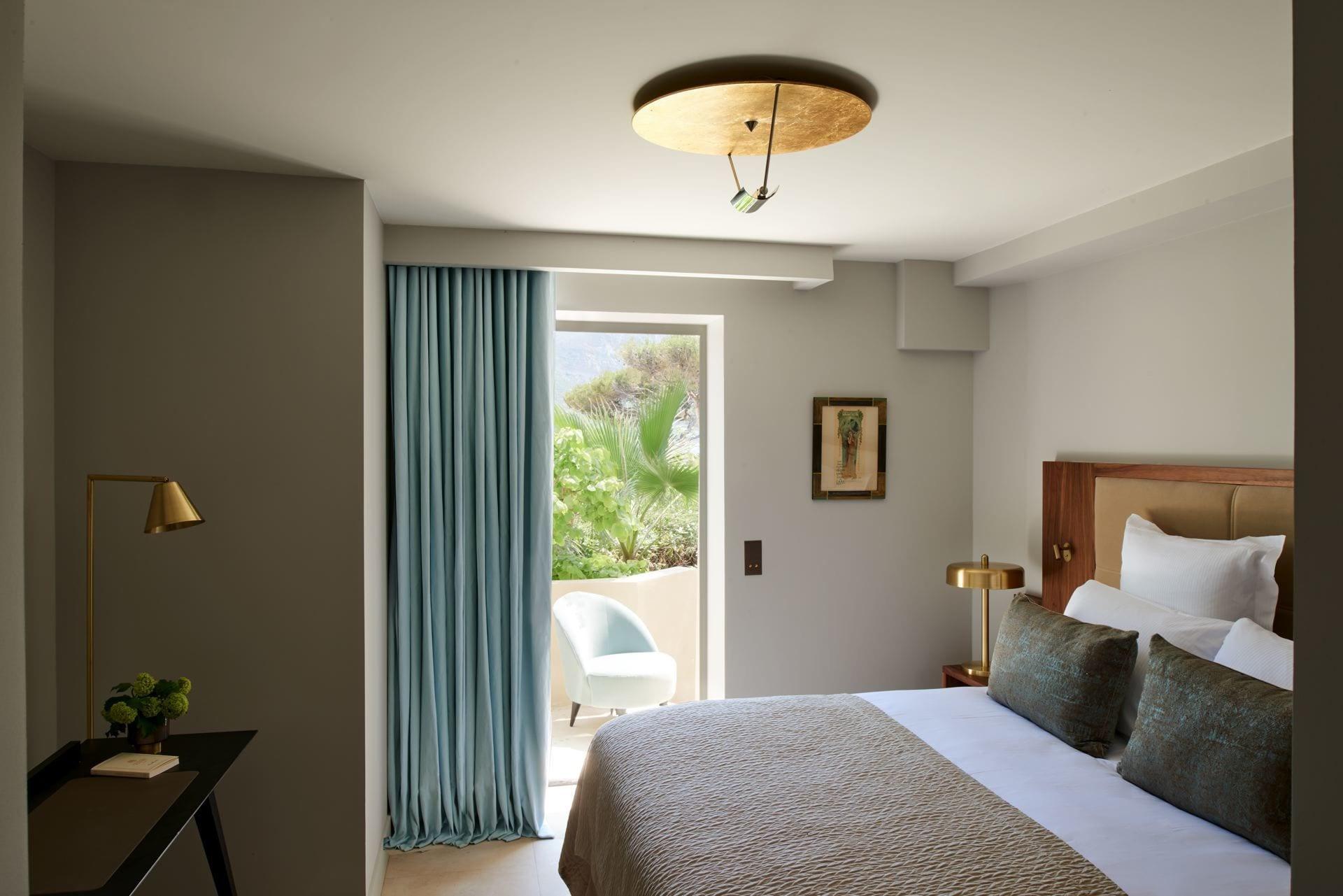 A bedroom with pale walls, brass ceiling fixture, and window with Mediterranean light