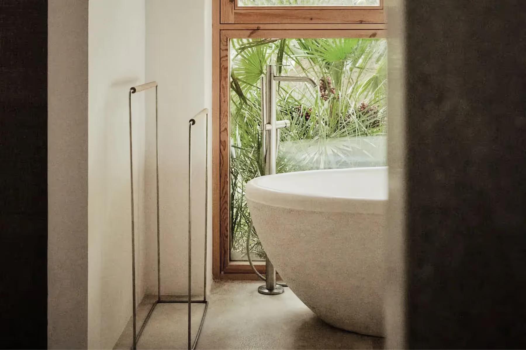 Es Racó d’Artà: freestanding sculptural bathtub positioned beside metal-frame window overlooking verdant landscape