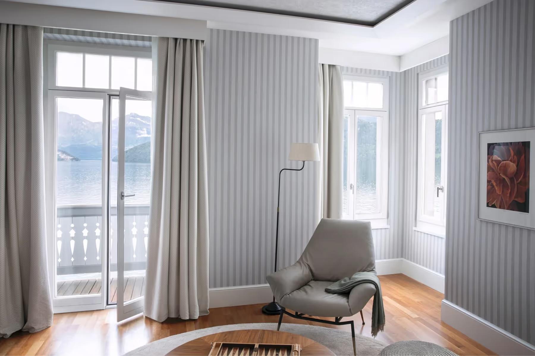 Bedroom corner with floor-to-ceiling windows, pale walls, lounge chair, and lake view