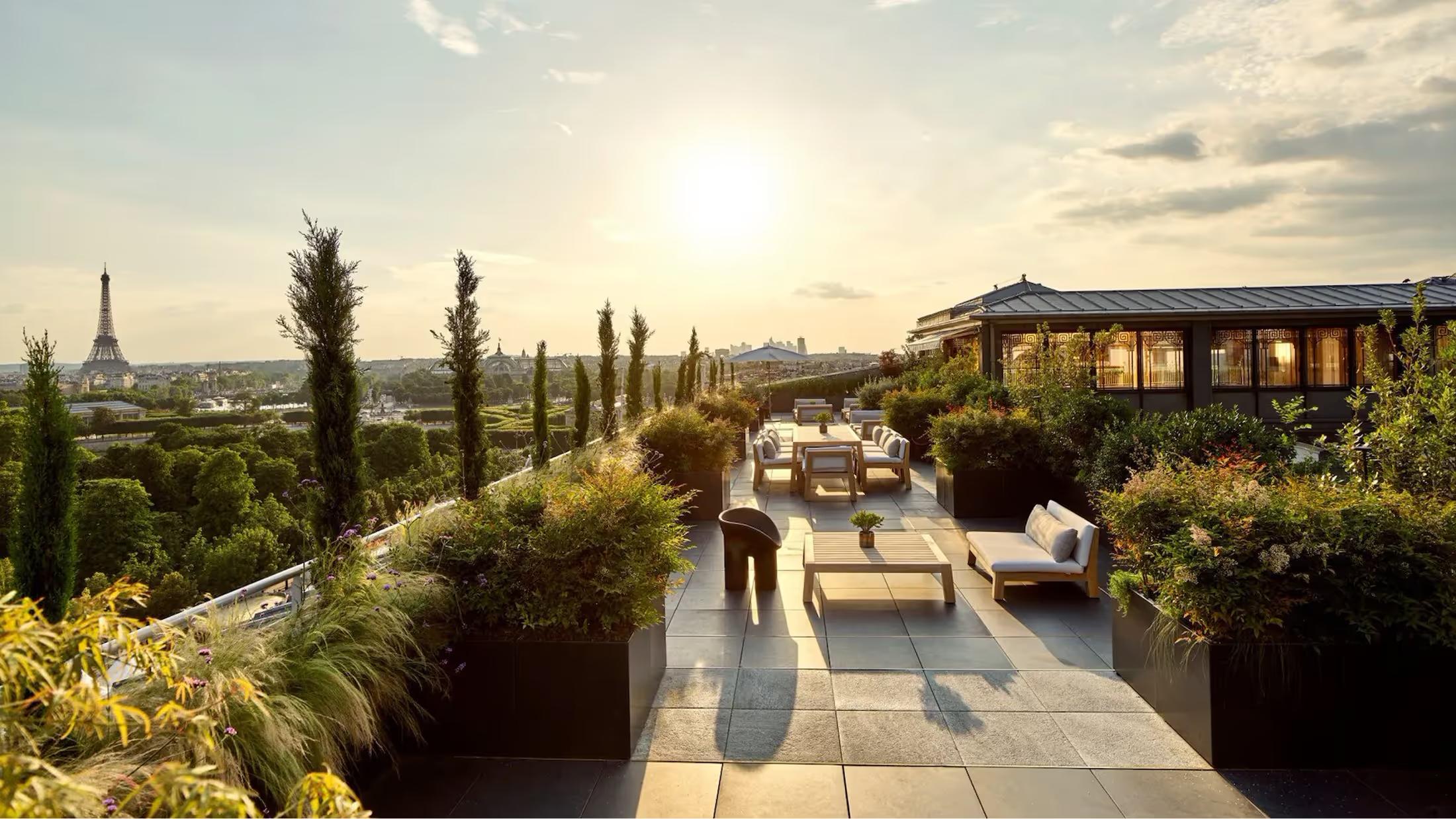 A terrace with a Paris skyline view, featuring cypress trees, loungers, and an umbrella at sunset