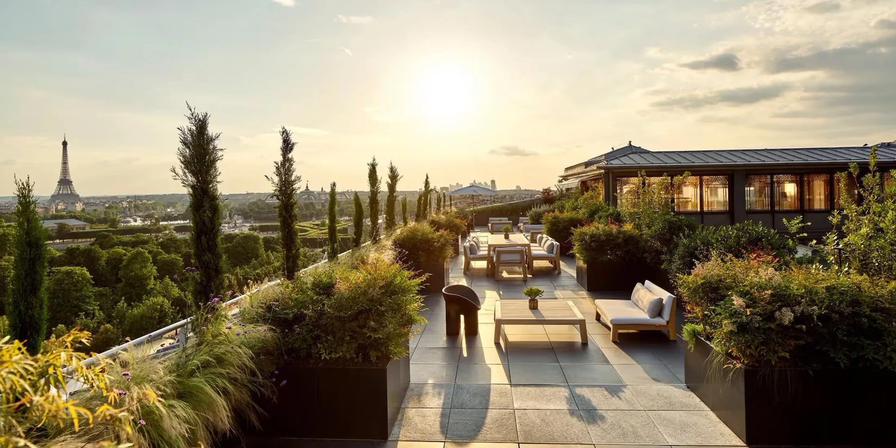 A terrace with a Paris skyline view, featuring cypress trees, loungers, and an umbrella at sunset