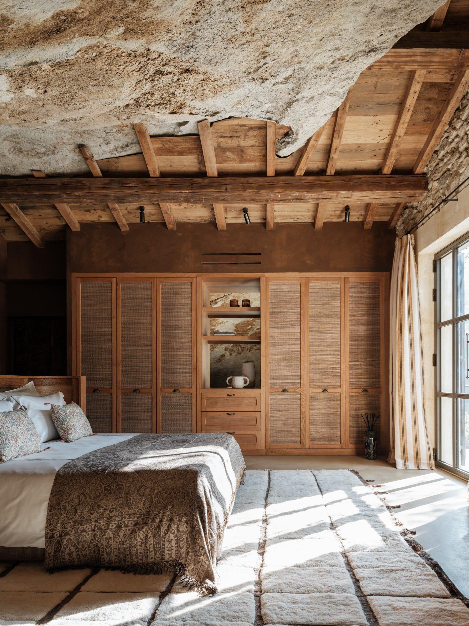 Rock-hewn bedroom with soaring raw limestone ceiling, wooden rafters, timber wardrobe, woven Berber rug