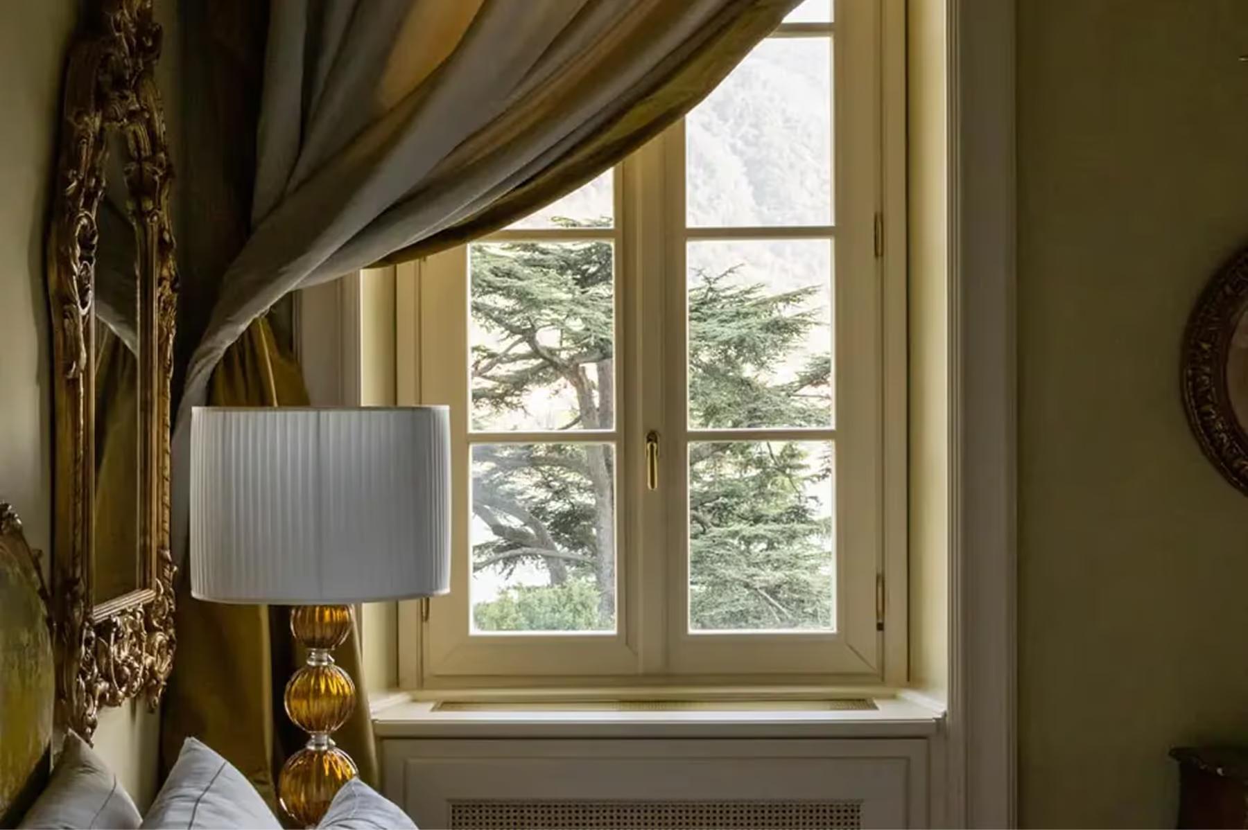 A window with gold damask curtains, lamp, and lake landscape visible through panes
