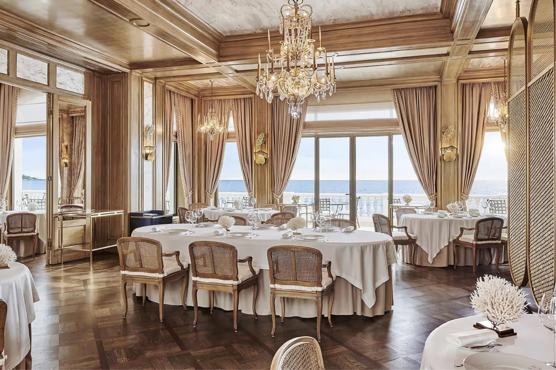 Grand salon dining space with round tables, high ceilings, and ocean backdrop